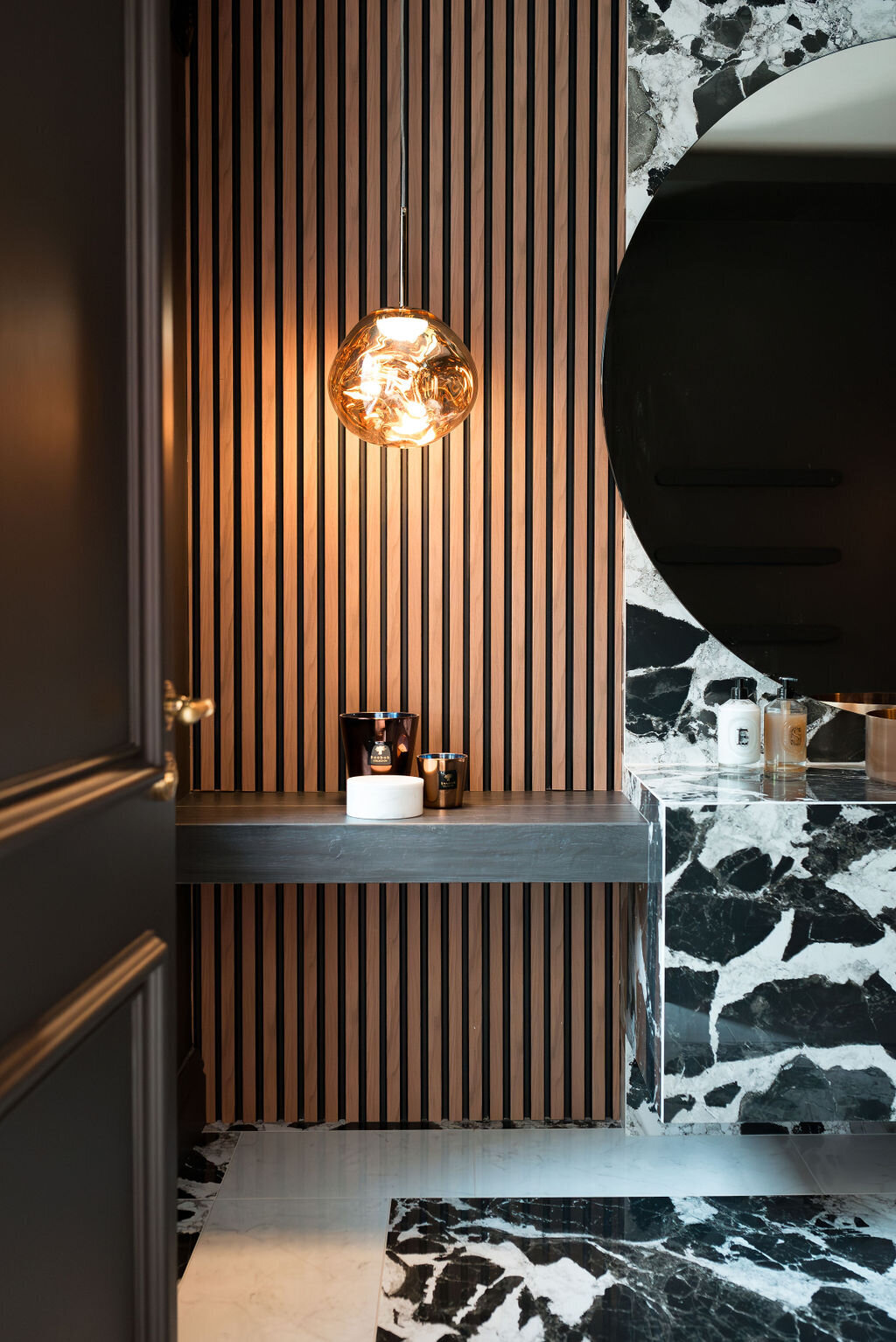 A modern bathroom with a large round mirror, black and white marble sink and countertop, wooden slatted wall, and pendant light fixture.