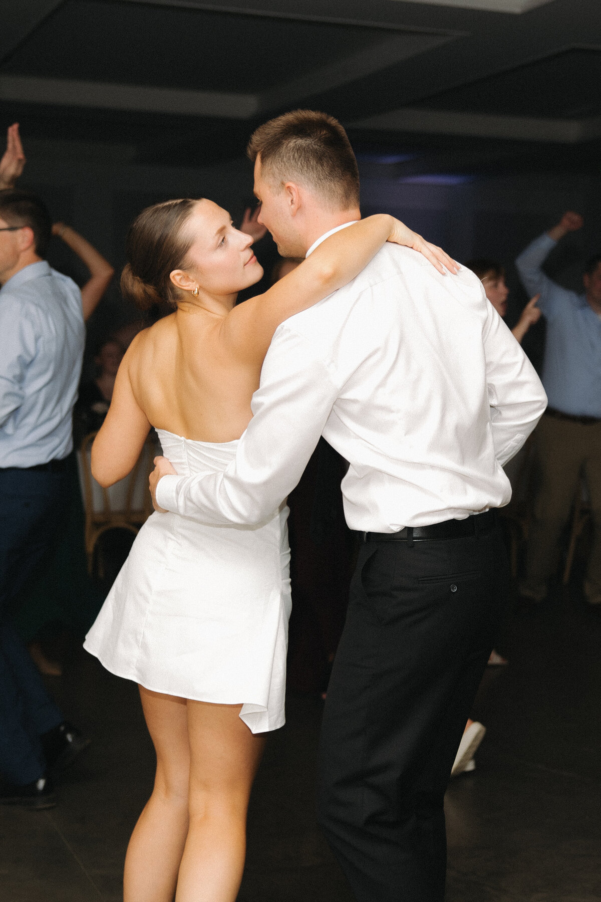 A bride in a short, strapless cocktail dress wraps her arm around her grooms shoulder and pulls him in so their noses are almost touching. The groom wraps his arm around her lower back. He is in a white dress shirt and black slacks. People dance behind them with their fists in the air. 
