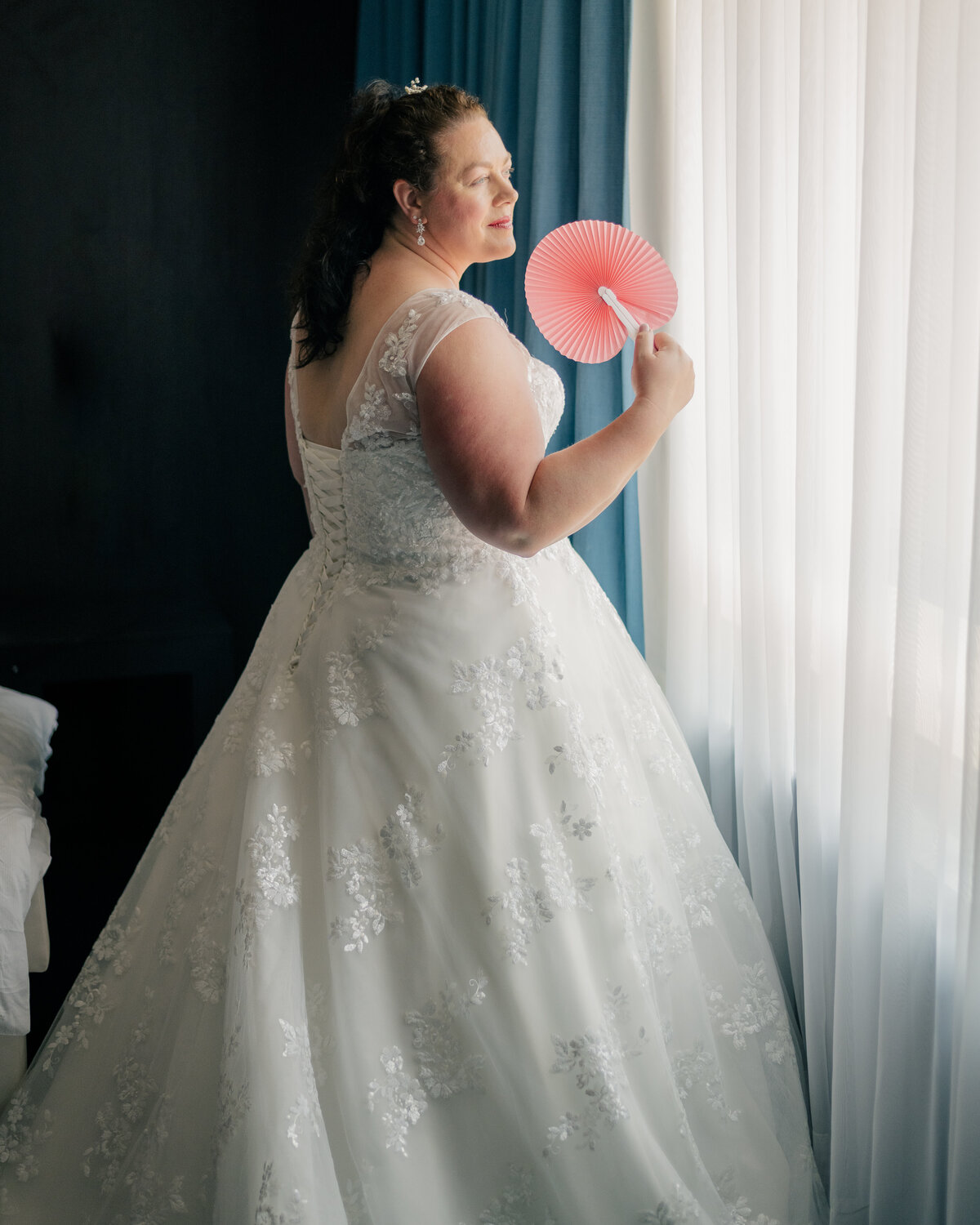 A bride stands in a hotel room before her wedding fanning her face at a window and looking outside