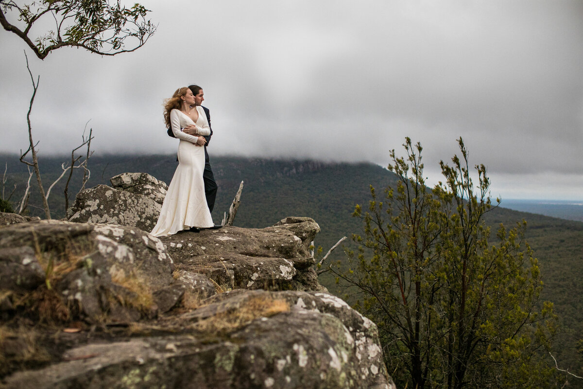 Bride and Groom standing on a mountain top. The wind is blowing in her hair and they are looking out to the view while hugging each other. The bride is a long sleeved wedding gown.