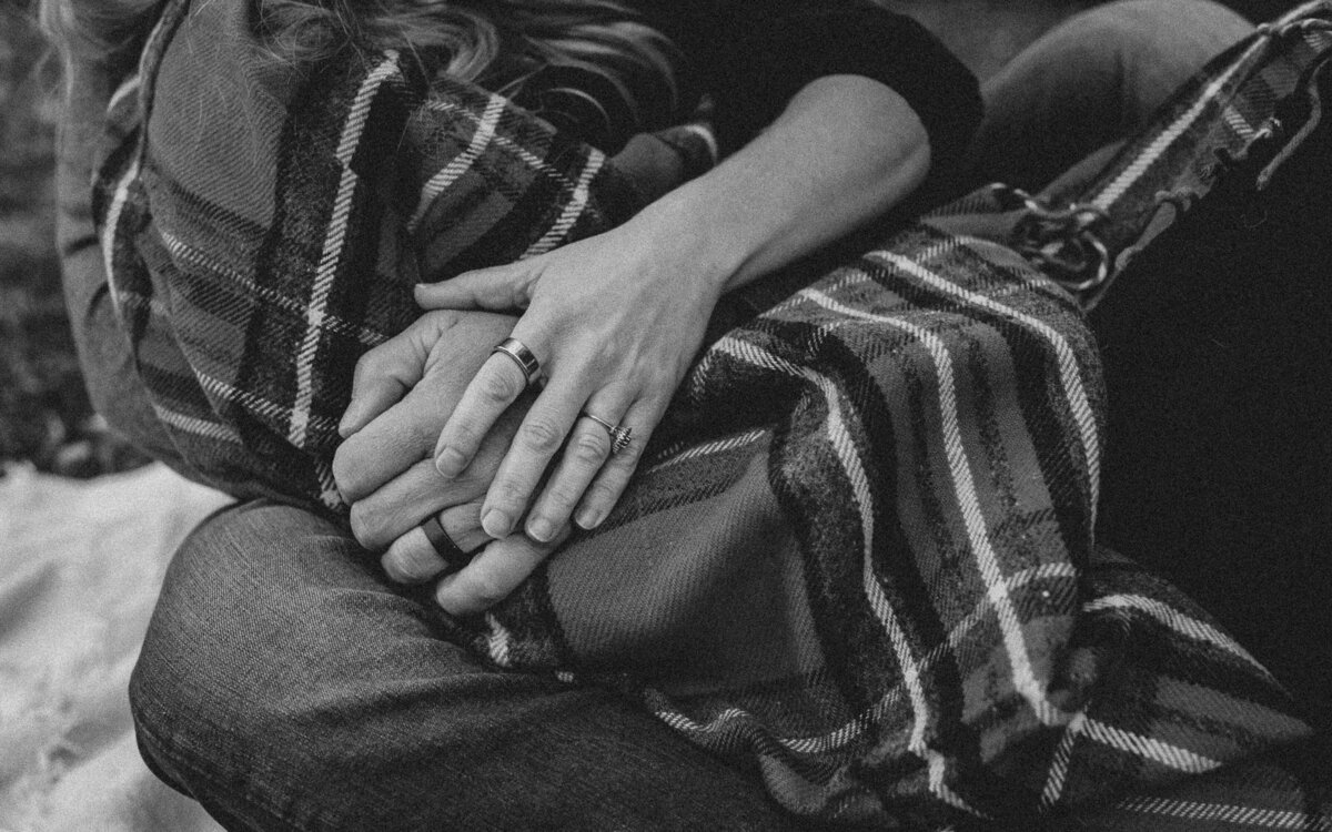 A black and white fine art image of a husband and wife's hands overlapping each other with a textured plaid blanket as the backdrop.