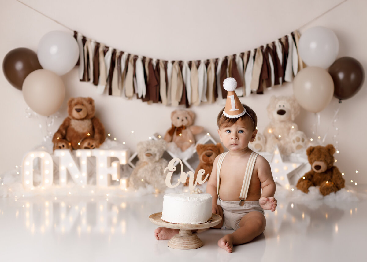 Neutral teddy bear cake smash in West Palm Beach in Wellington Florida portrait studio. Baby boy is wearing khaki Shortys with suspenders. The background is a soft cream with plush teddy bears and lights.