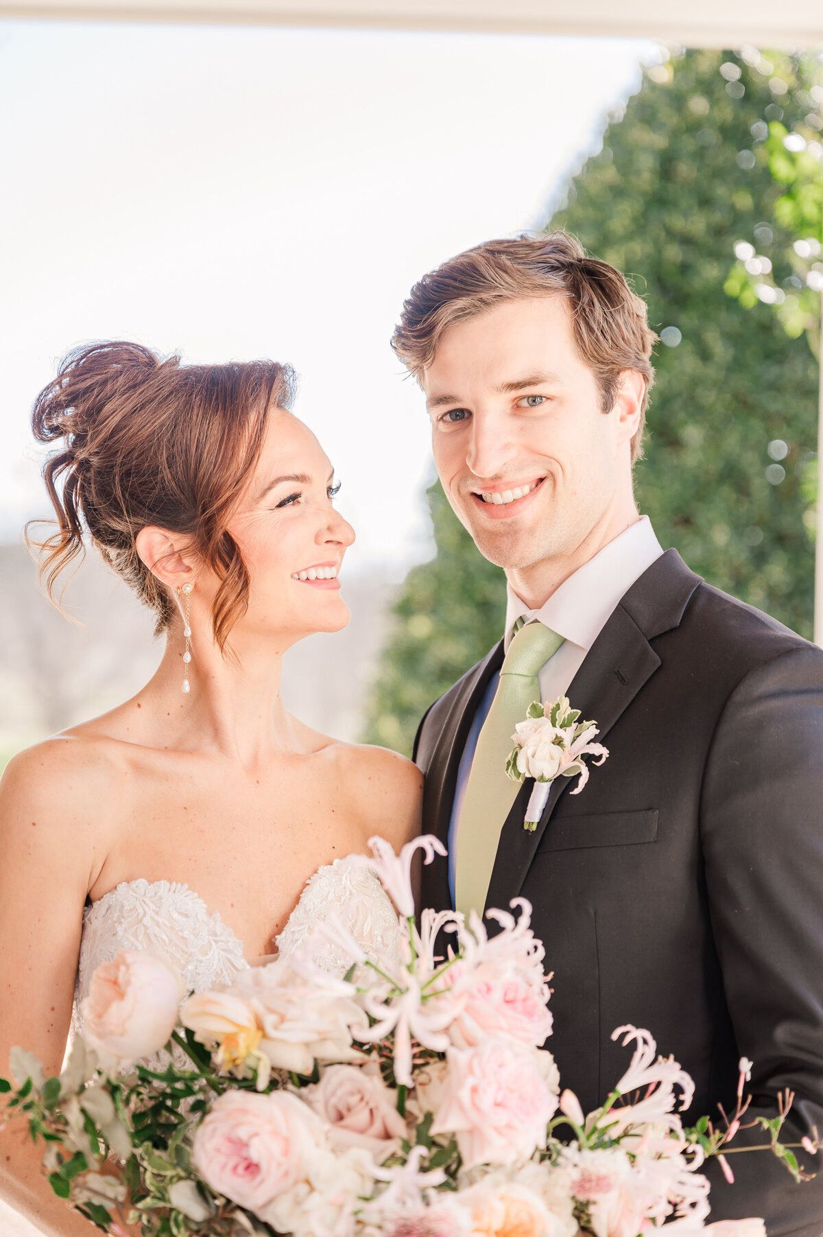 A bride in a stunning white lace wedding gown holding a pastel wedding bouquet smiling at her husband in a black tux and green tie at Donovan Manor by JoLynn Photography, a Raleigh wedding photographer