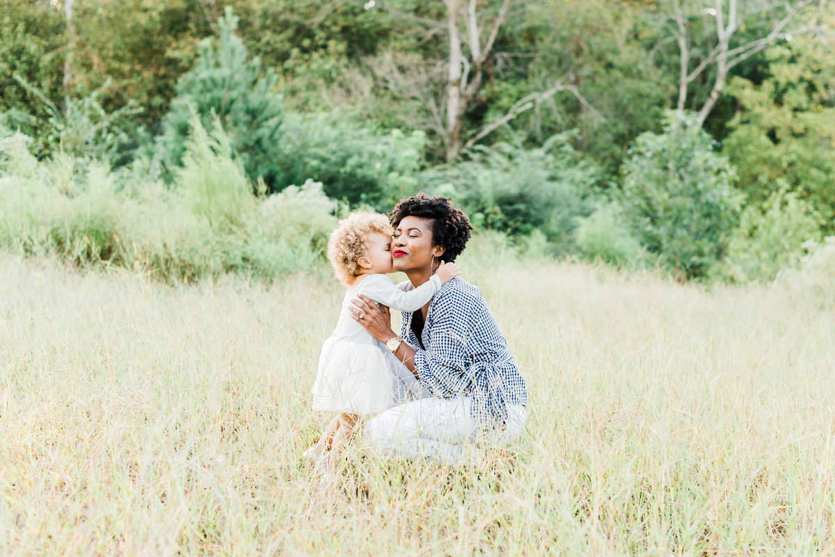 Mom bends down in a field for her daughter to kiss her on the cheek during a Raleigh family portrait session. Photographed by Raleigh family photographer A.J. Dunlap Photography.