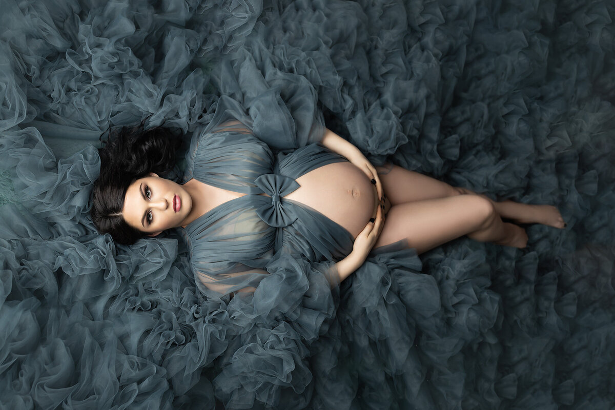 Woman lying on a bed of teal tulle, hands gently resting on her belly, wearing a flowing sheer gown with a bow detail. Maternity photography by Lindsay Walden Photography.