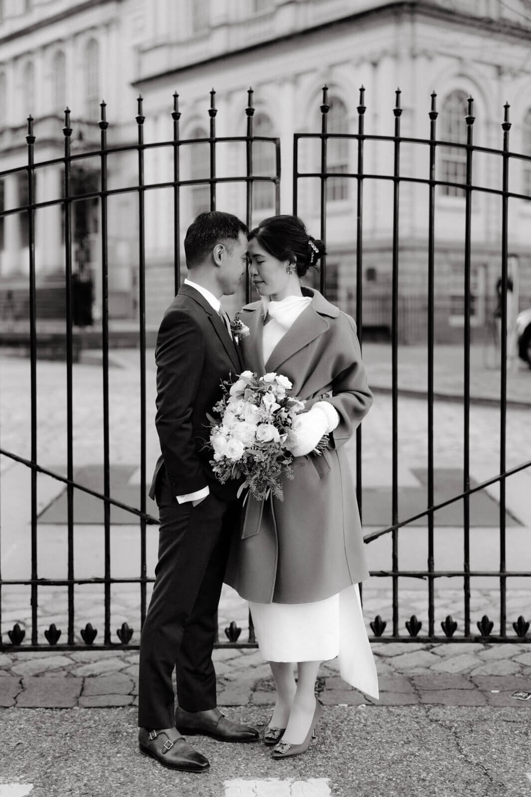 Black and white photo of the bride with flower bouquet, and the groom, standing in front of the gate of an old building.