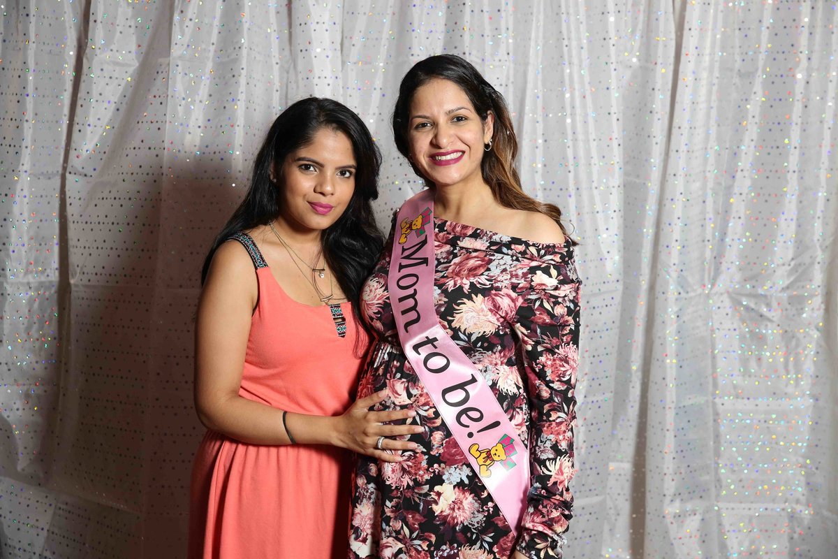 A pregnant lady poses with a female friend in front of a mild curtain. Photobooth by Ross Photography, Trinidad, W.I..