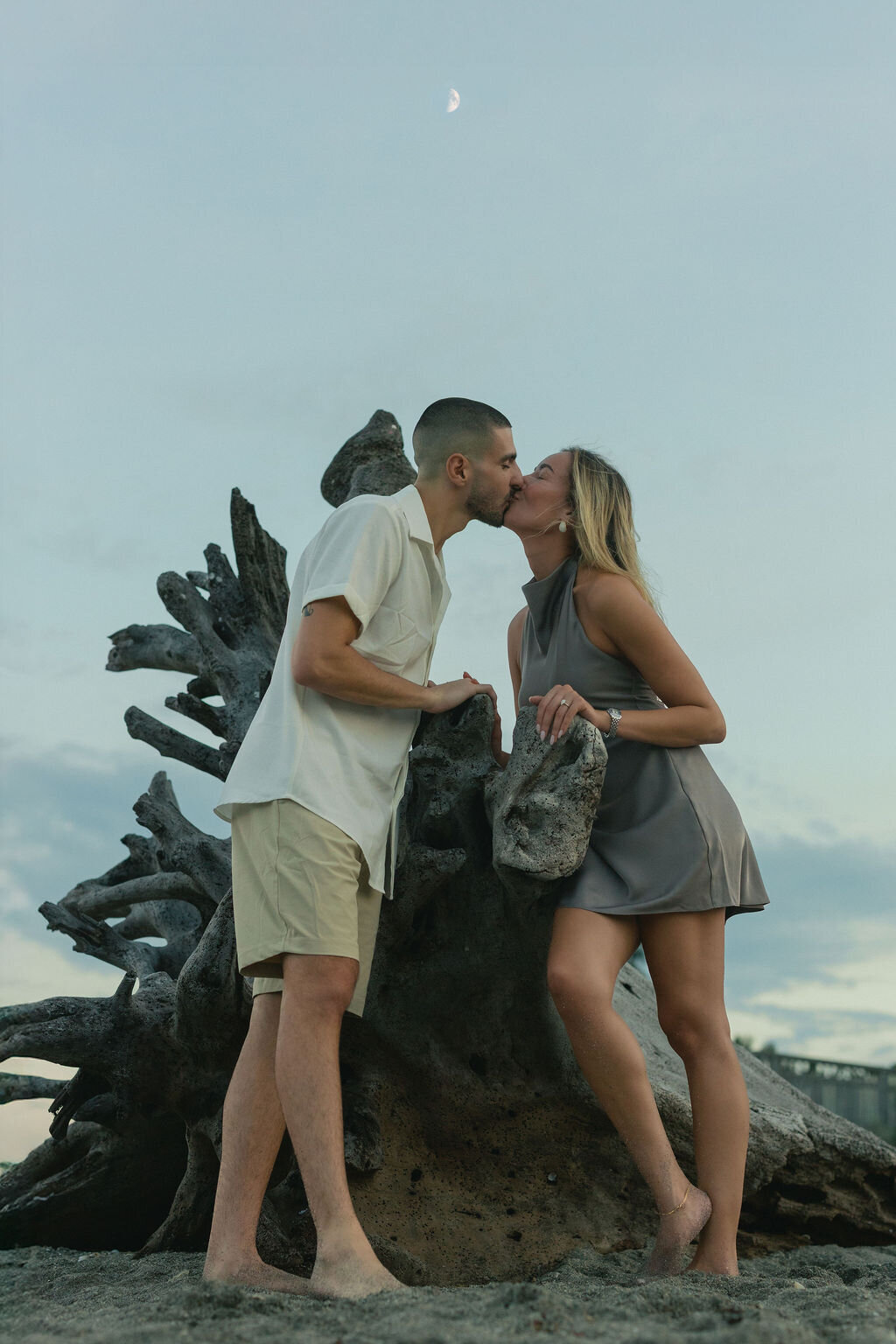 A couple kisses by a washed up tree at the blowing rocks preserve beach in Jupiter, FL with the moon high in the sky at blue hour.