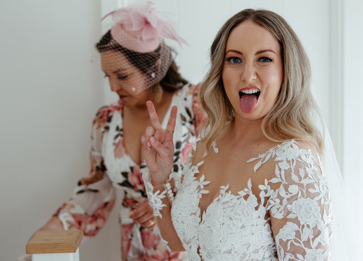 A bride in a lace wedding dress making a playful peace sign, with a bridesmaid assisting in the background