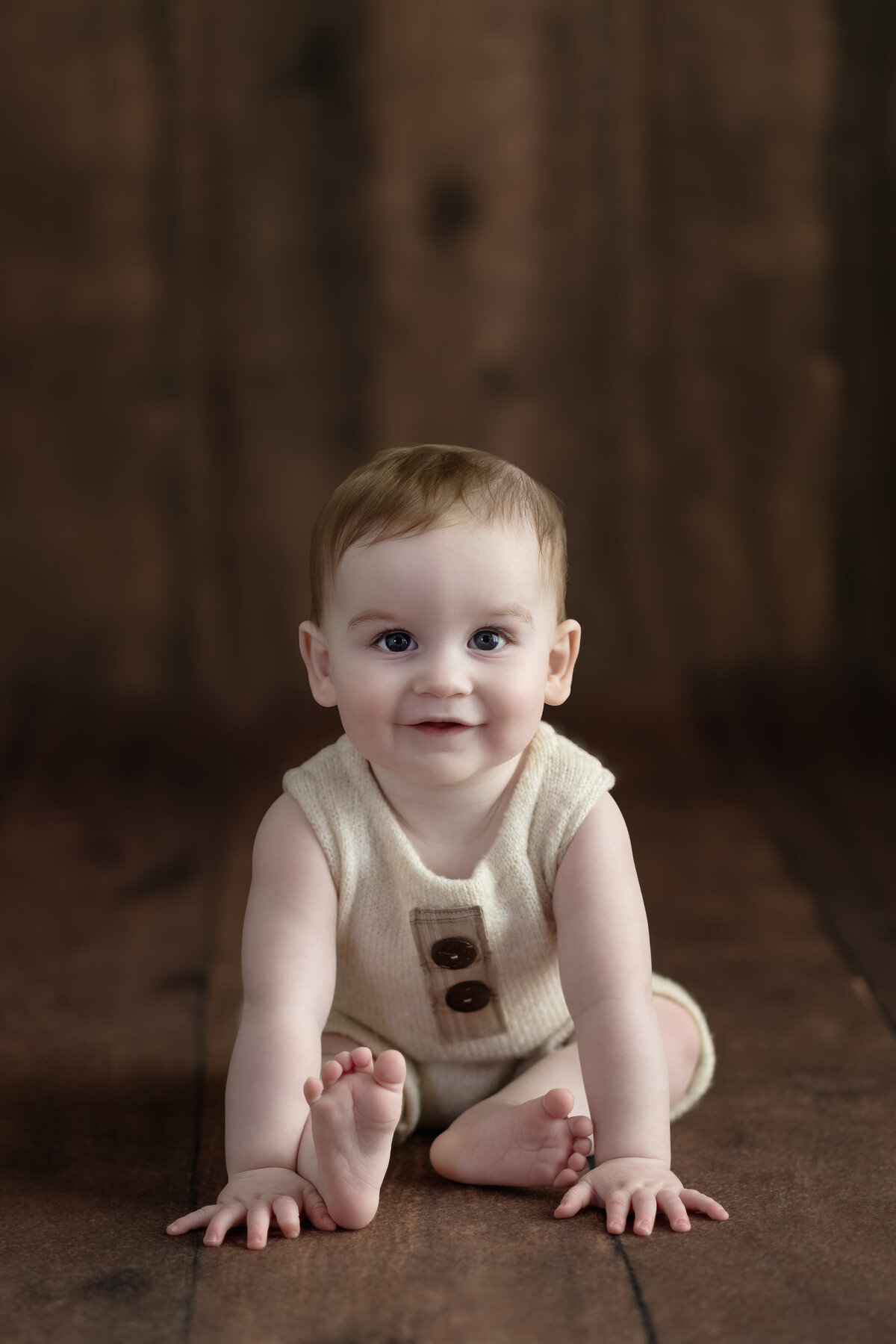 Milestone sitter photo of baby boy in a simple romper on warm wood—timeless textures, tender gaze.