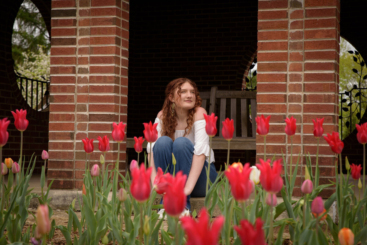 Green Bay East High School senior girl wearing a white sleeveless shirt and bell bottom jeans sitting behind a tulip garden in the red brick building at the Green Bay Botanical Gardens in Green Bay, Wisconsin.