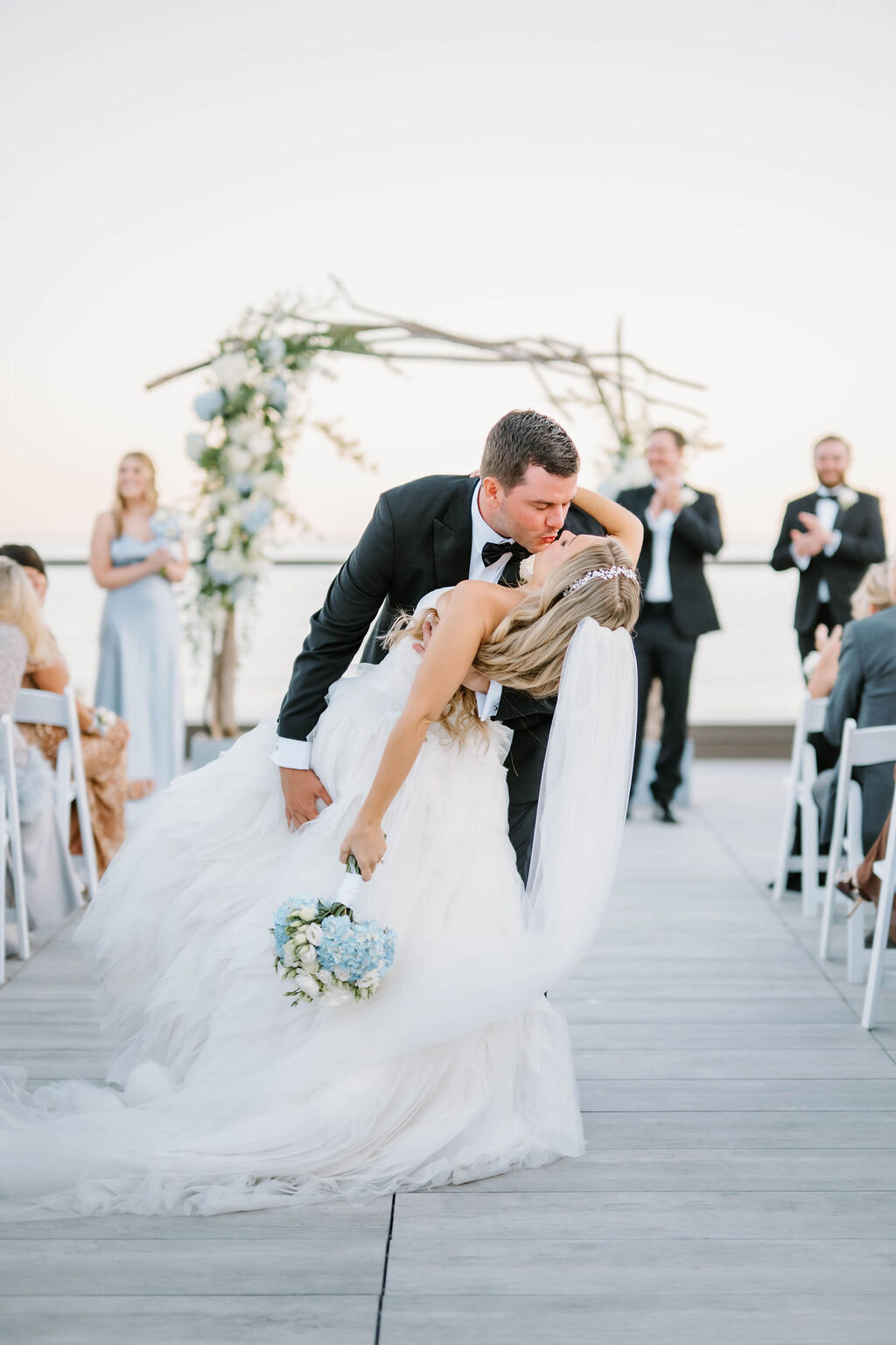 Rhode Island Wedding Photographers | A groom dips the bride for a kiss in front of an outdoor altar adorned with flowers. Guests stand and clap.
