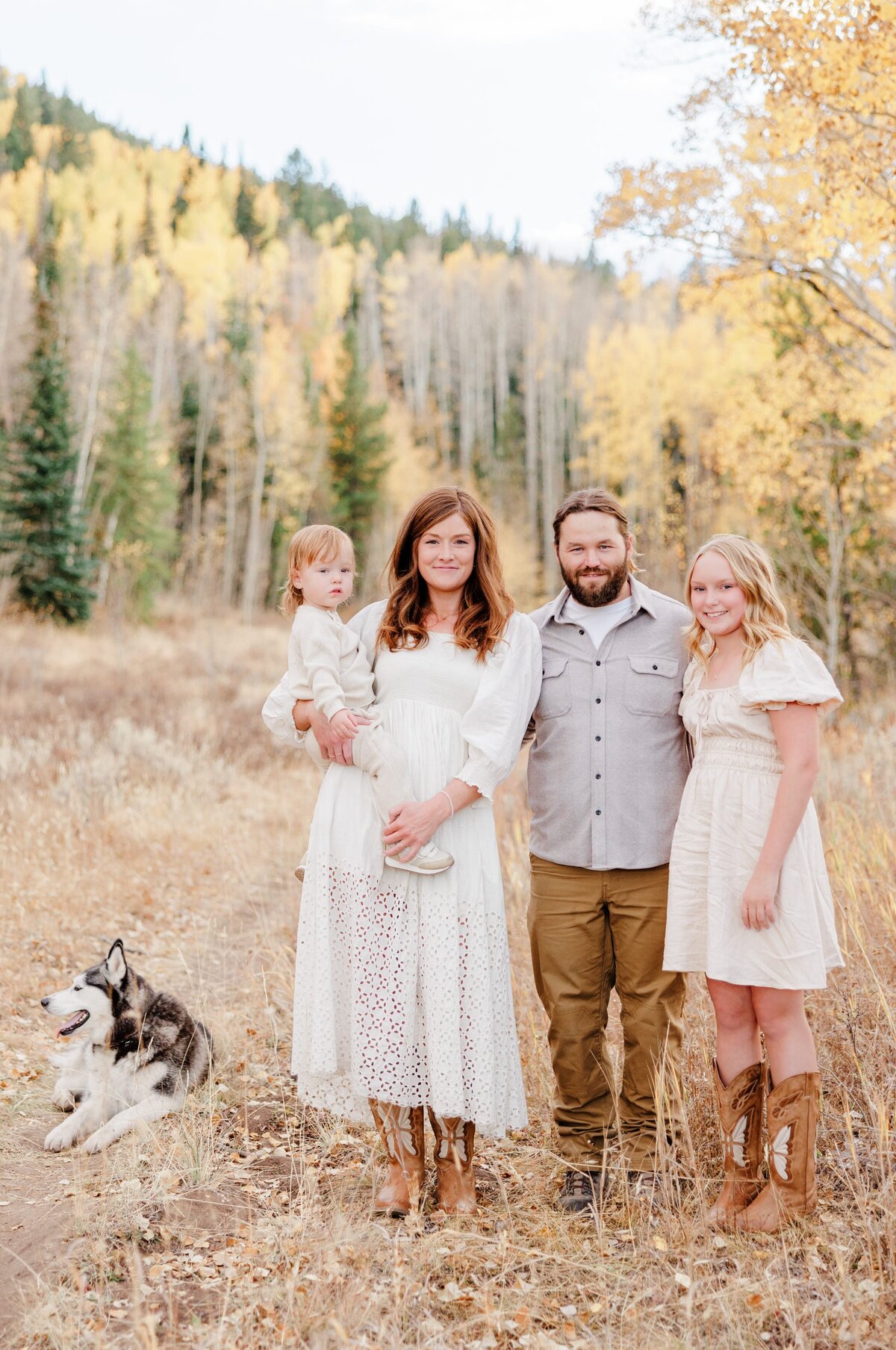 A husky lays behind its family standing in a trail smiling in fall
