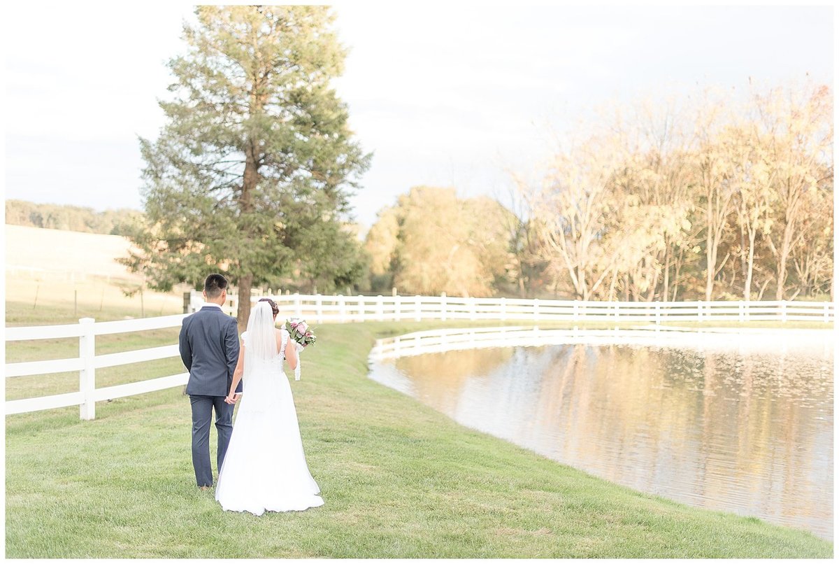 Bride and Groom holding hands and walking with backs toward camera beside white fence and large pond.