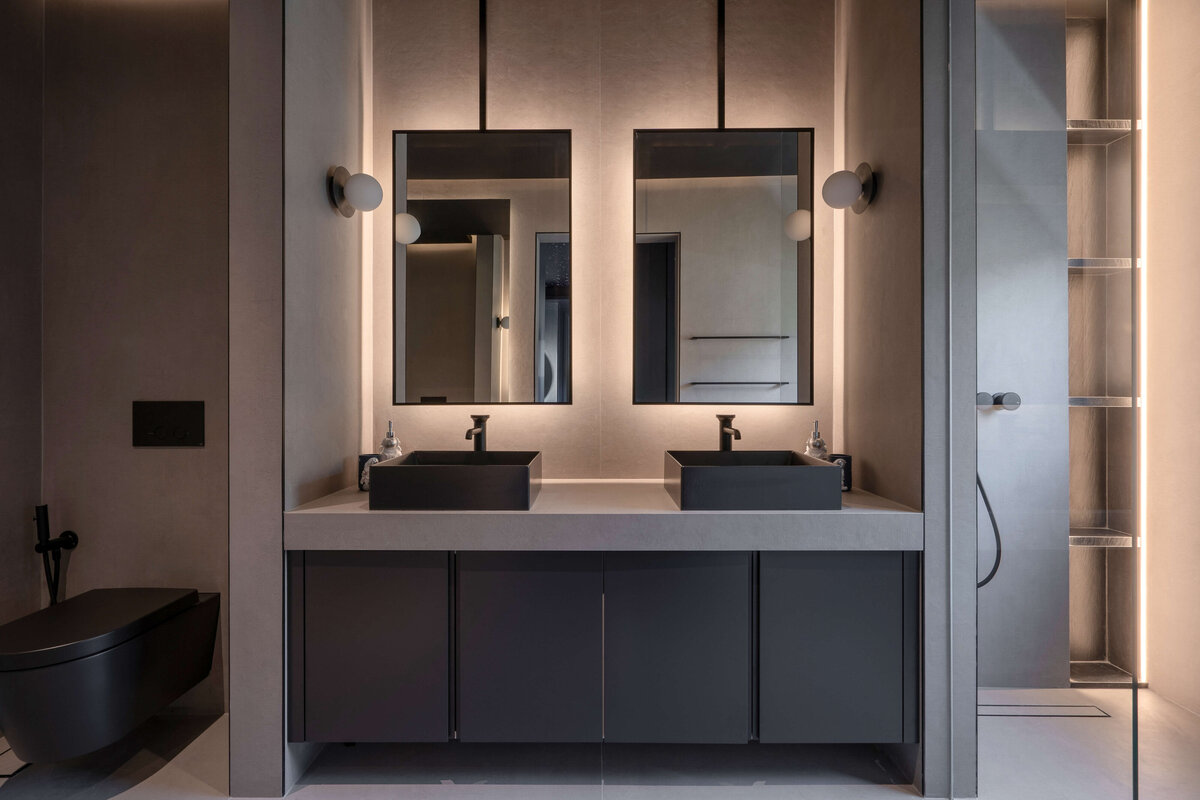 Sleek grey bathroom with dark cabinets, light flooring, and double black sinks.. To the left of the sinks is a contemporary black toilet. To the right of the sinks is a walk-in shower.