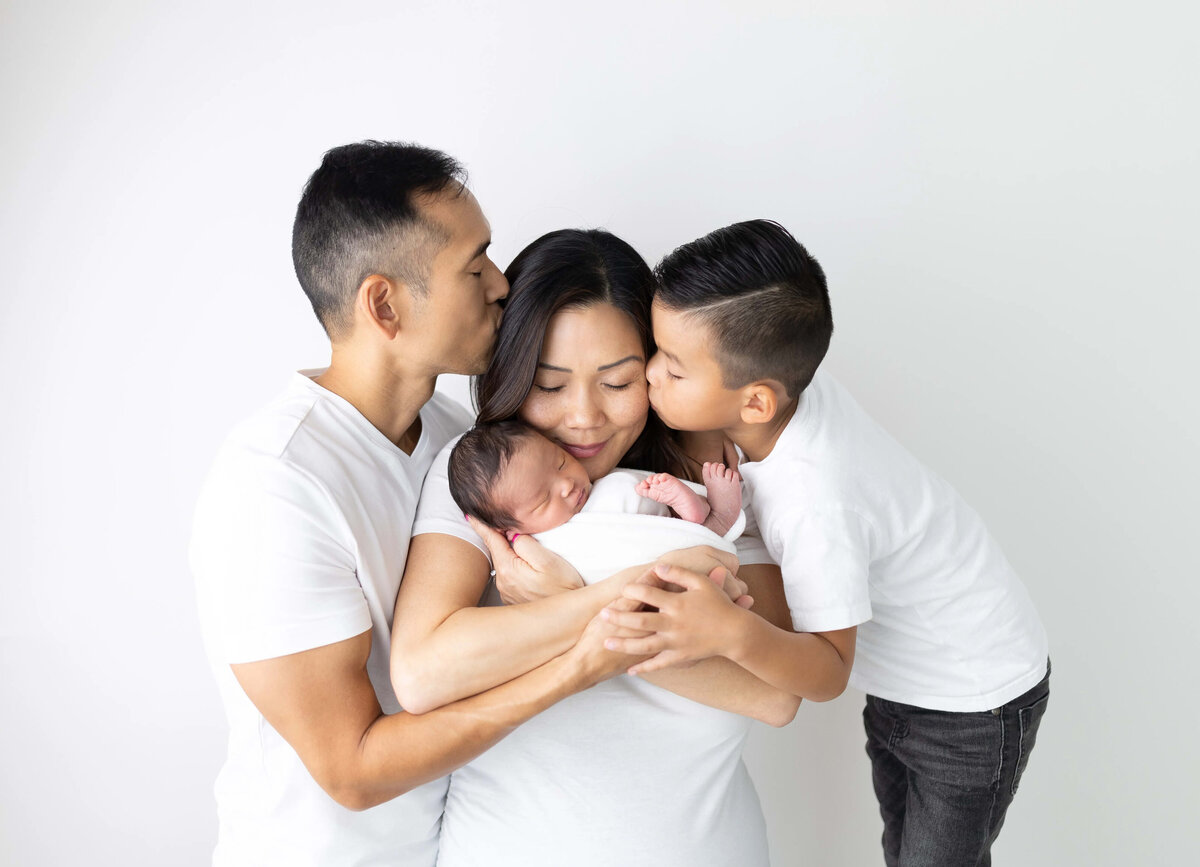 Mom holding a newborn while dad and big brother kiss her - family newborn session in white studio 