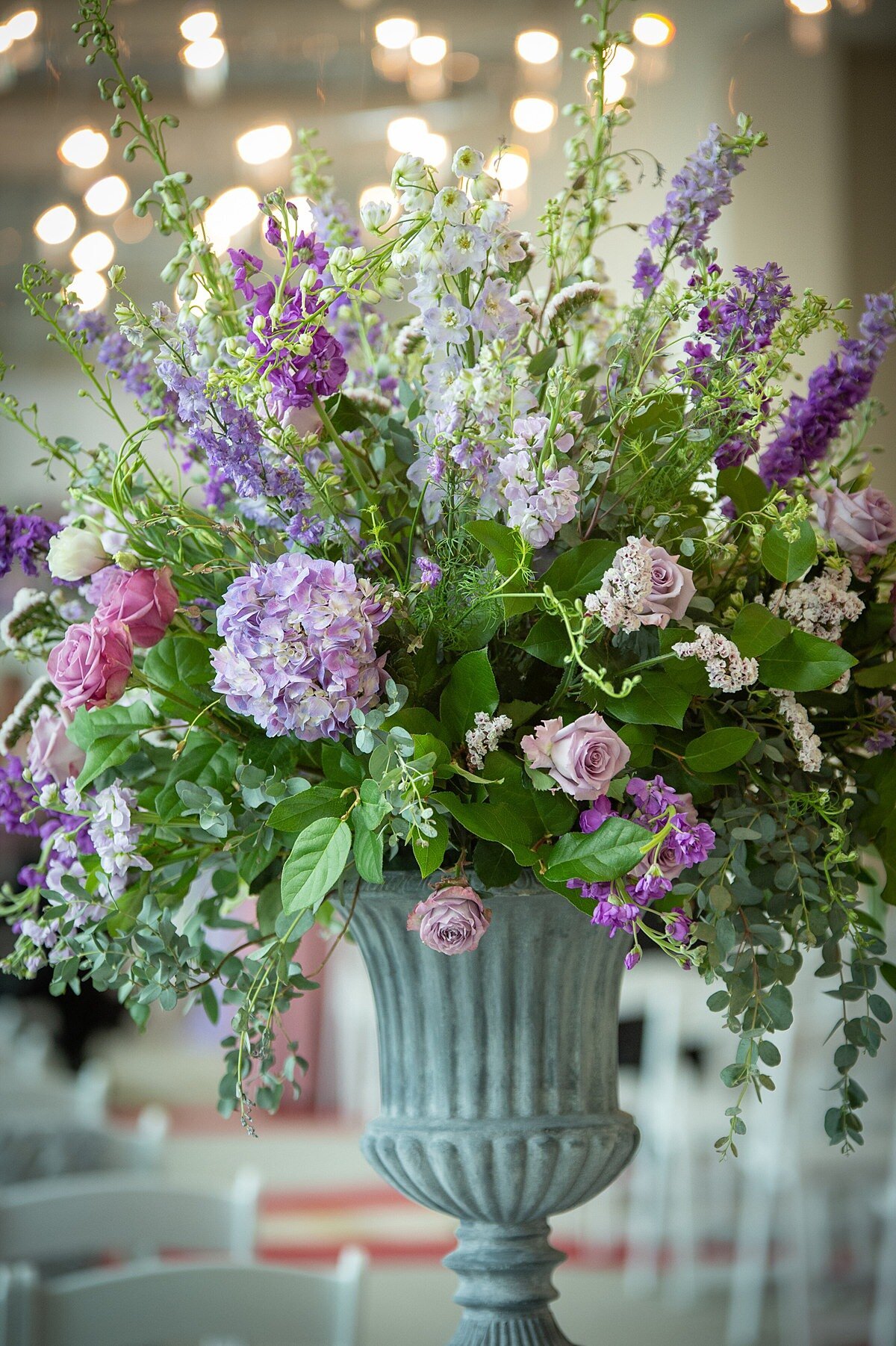A large floral arrangement in a grey stone urn at Noah Liff Opera Center features pink roses, light purple roses, purple hydrangea, purple stock eucalyptus and greenery at a wedding with white garden chairs.