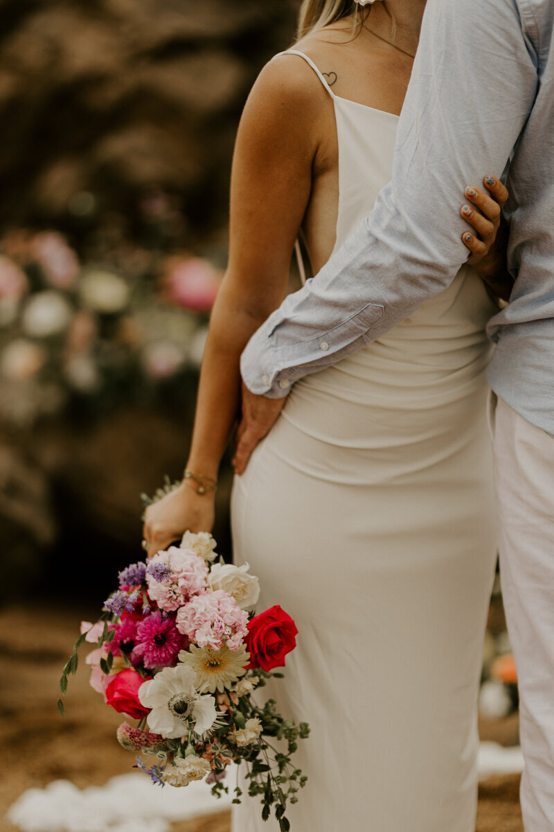 Femme en robe fluide blanche tenant un bouquet coloré, la main de son chéri posé sur sa hanche. Shooting photo en Vendée.