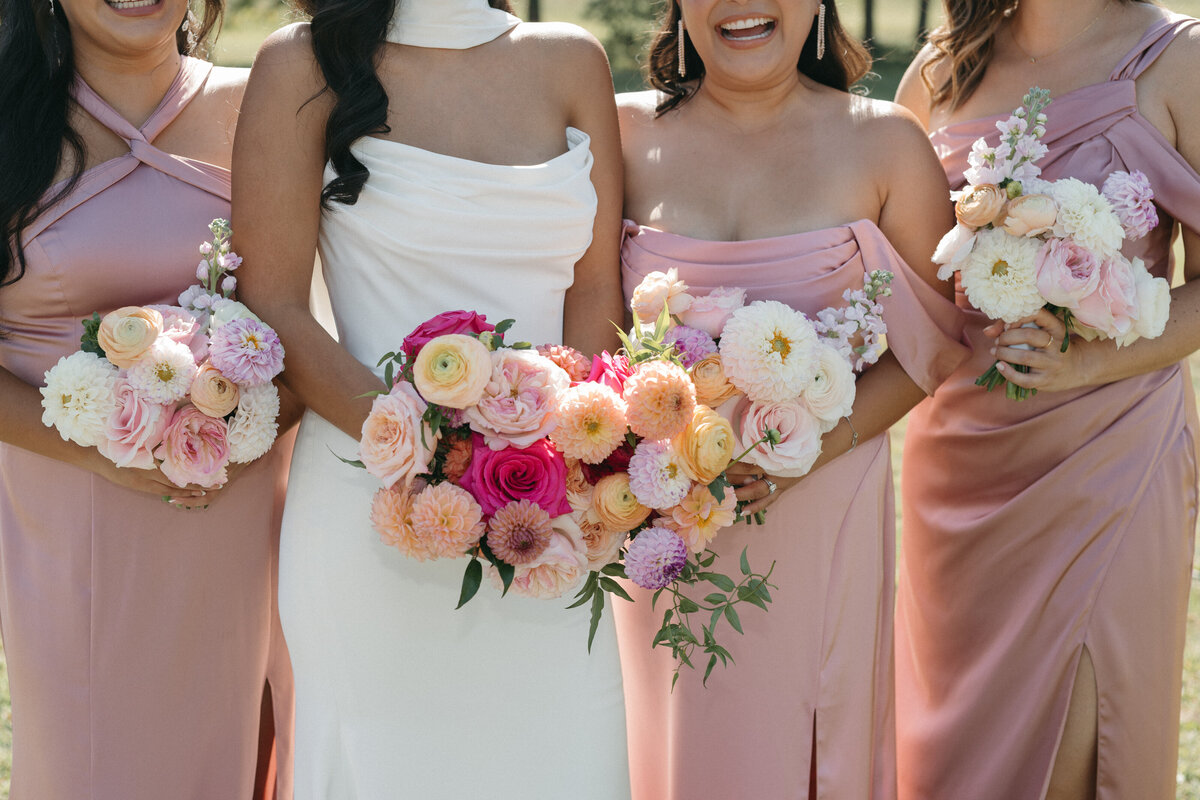 Bride and bridesmaids holding vibrant garden-style bouquets with dahlias, roses, and ranunculus in peach and pink tones, styled with blush satin dresses for a romantic outdoor wedding.
