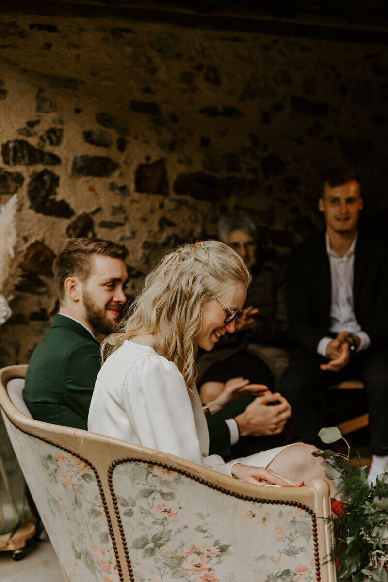 Mariés de profil assis dans un canapé au style ancien et souriant capturée par Laura, photographe de mariage en Vendée.