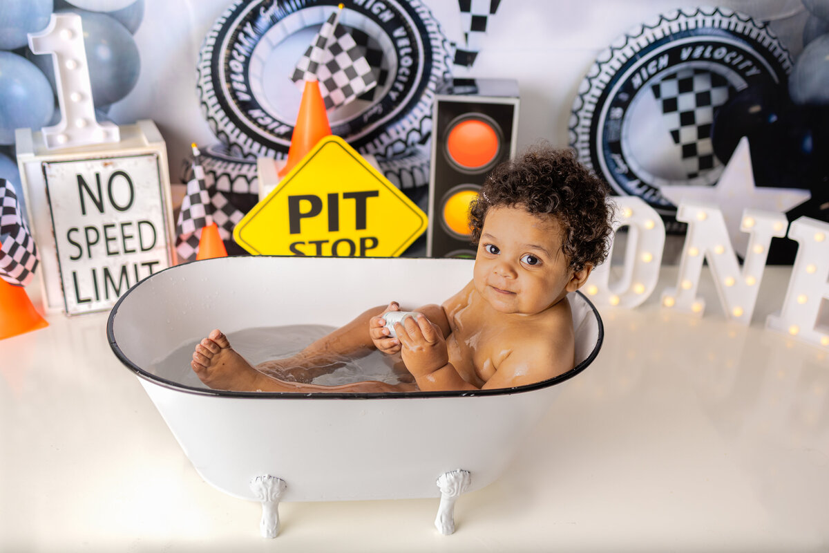 Baby boy relaxing in mini bathtub during race car themed cake smash session with pit stop decor and checkered flags in Waldwick NJ