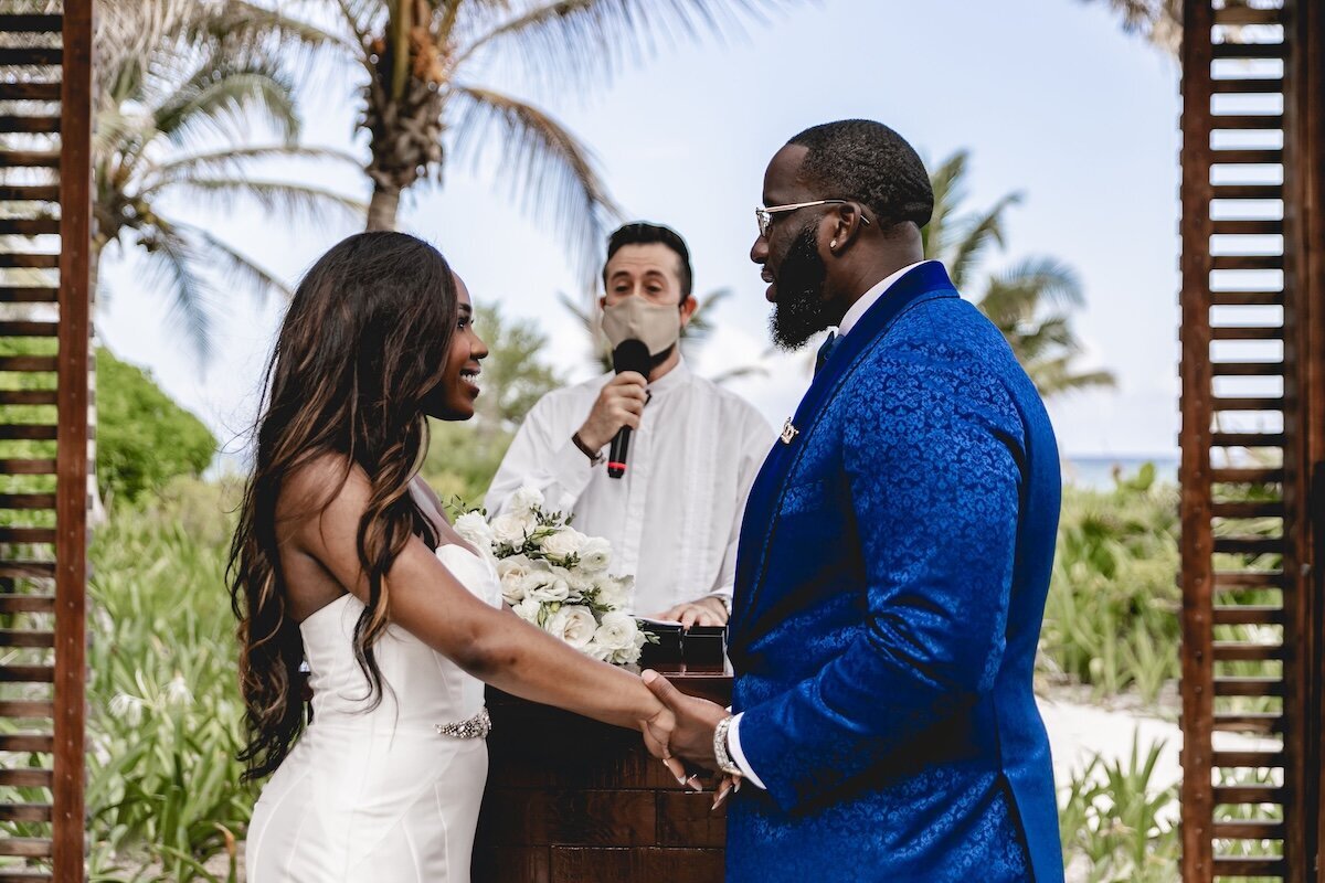 A bride and groom hold hands during their outdoor wedding ceremony. The bride is wearing a strapless white gown, and the groom is dressed in a bright blue suit. They are standing in front of a tropical backdrop with palm trees, with the officiant standing behind them.