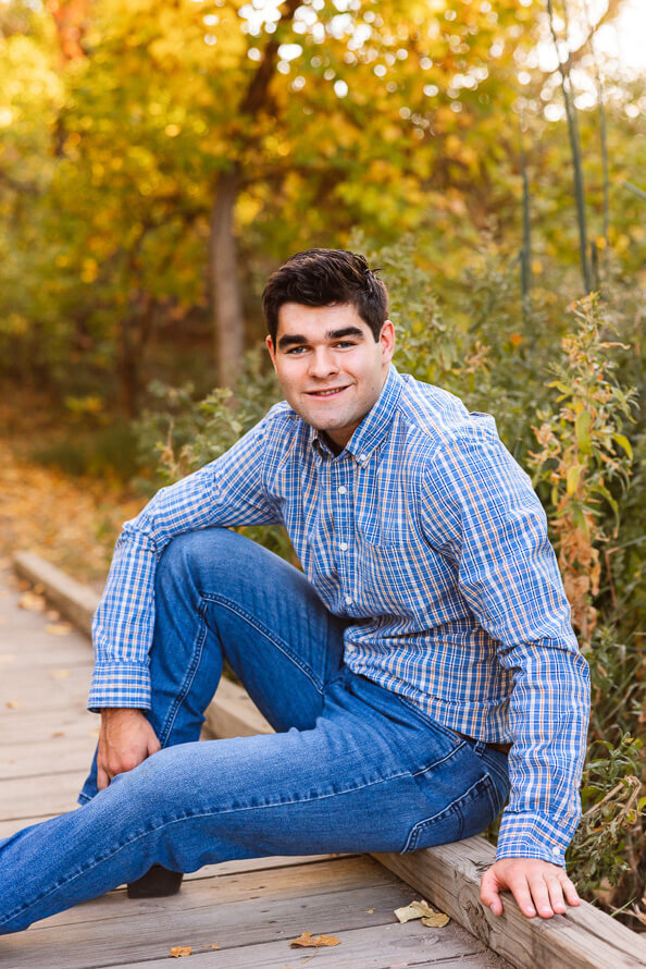Boy in blue jeans and a blue shirt sits on a bridge for his senior photos.