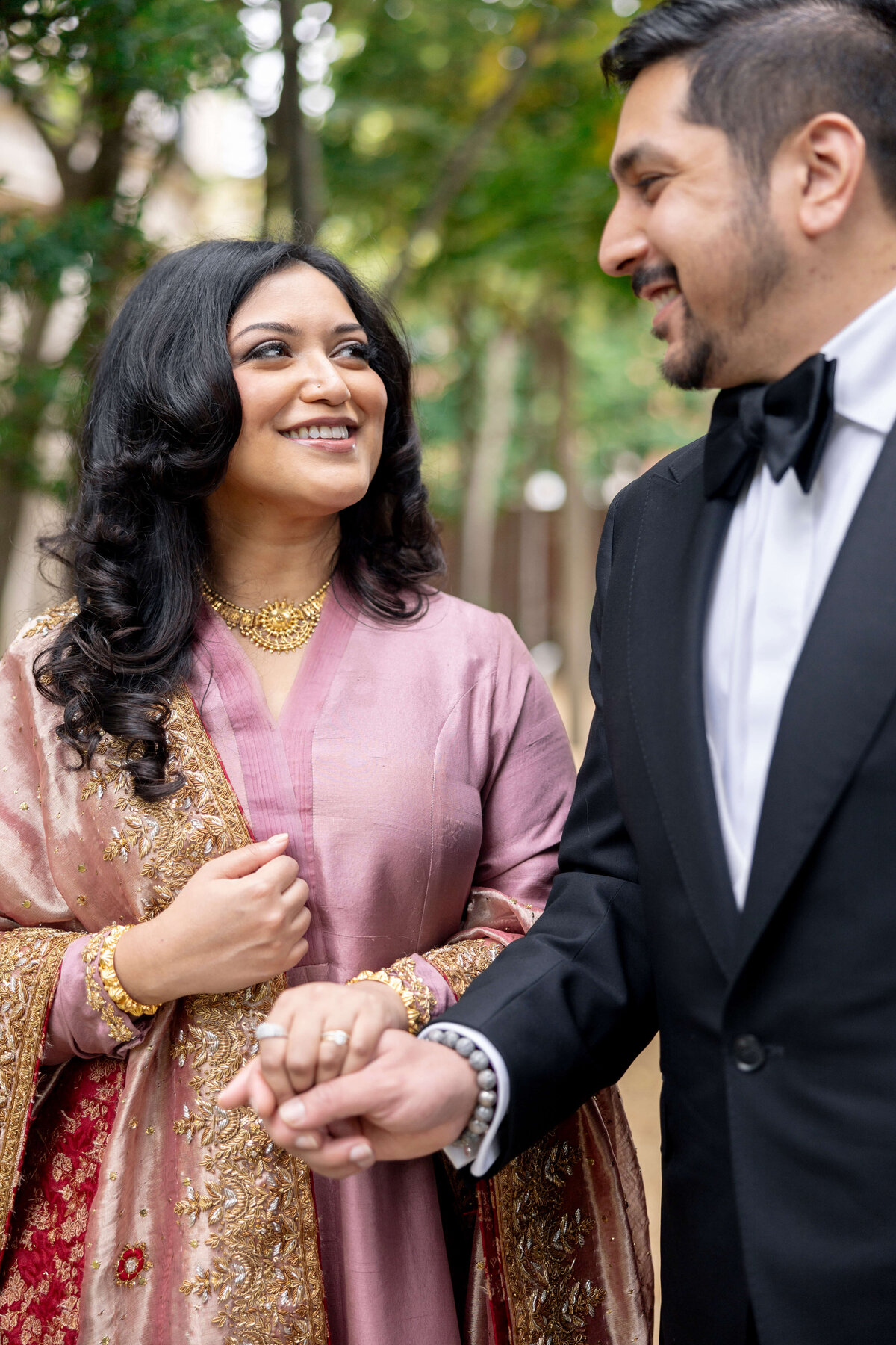 A couple holds hands and smiles at each other outdoors. The woman is wearing a pink traditional outfit with gold jewelry, and the man is in a black tuxedo. Trees are blurred in the background.