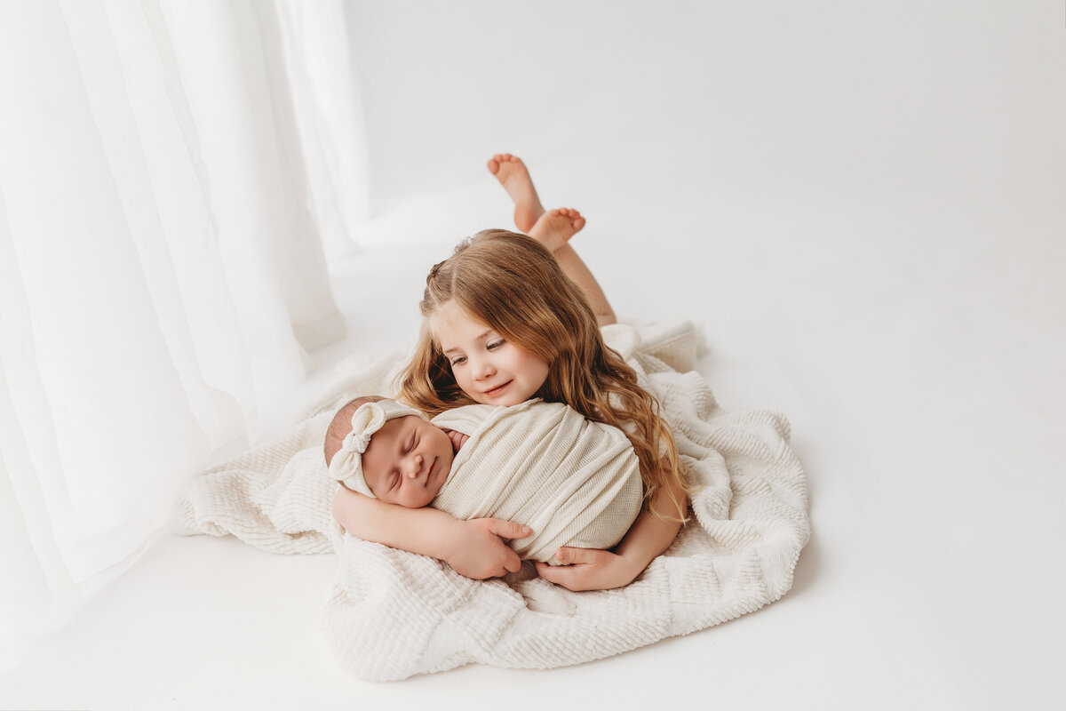Baby smiling with big sister on white backdrop for natural newborn session at phoenix newborn studio