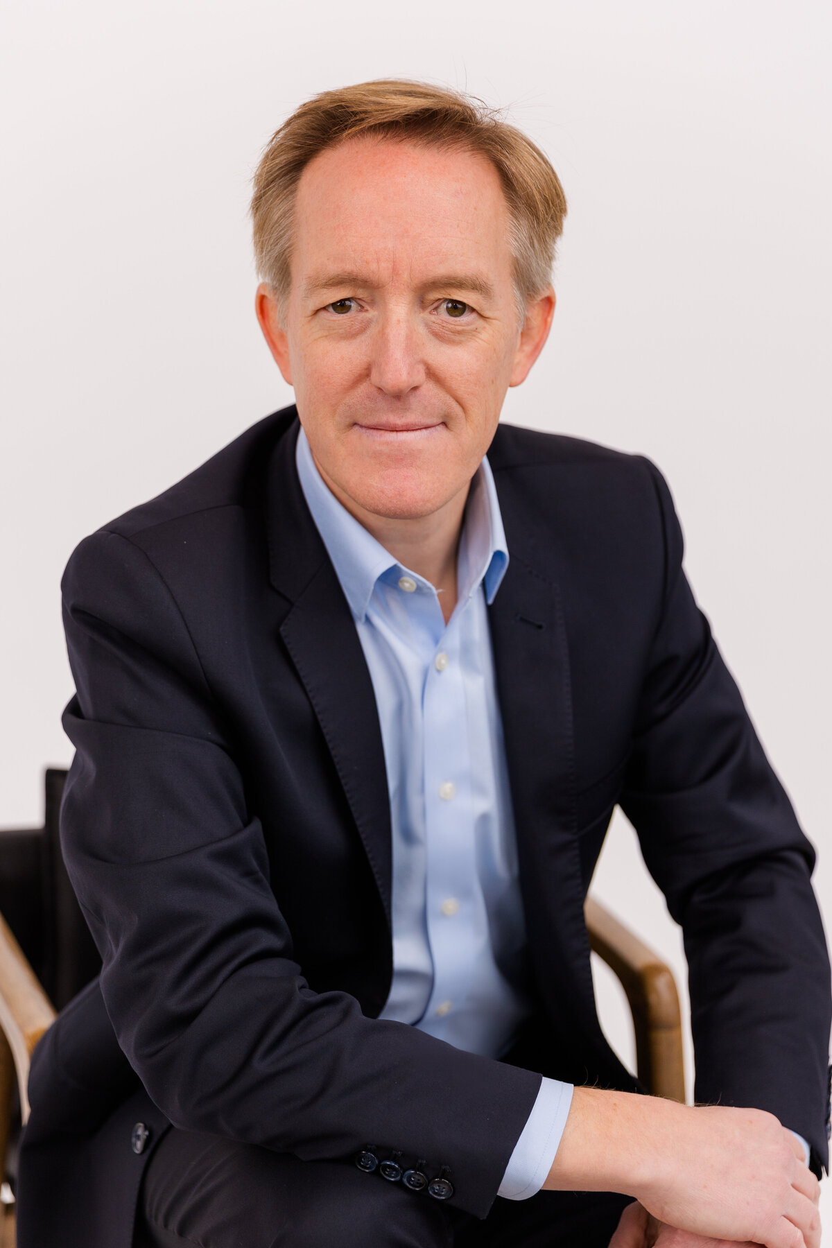 man sitting on a chair in front of white background wearing corporate outfit light blue shirt and dark jacket during headshot photoshoot with Laure Photography
