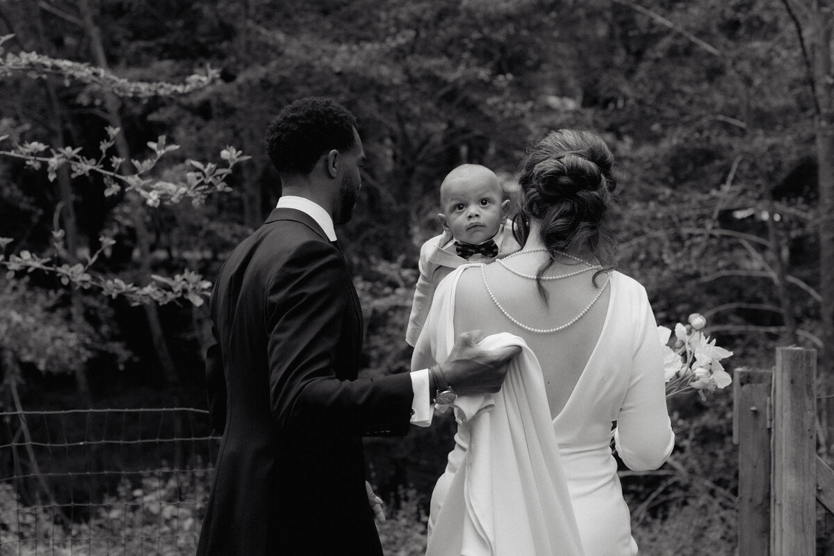 A timeless black and white wedding portrait featuring a bride, groom, and their baby in an intimate moment. The groom gently holds the bride’s gown as they walk, with the baby gazing toward the camera. We specialize in capturing heartfelt, documentary-style wedding photography that tells authentic love stories with a timeless aesthetic.