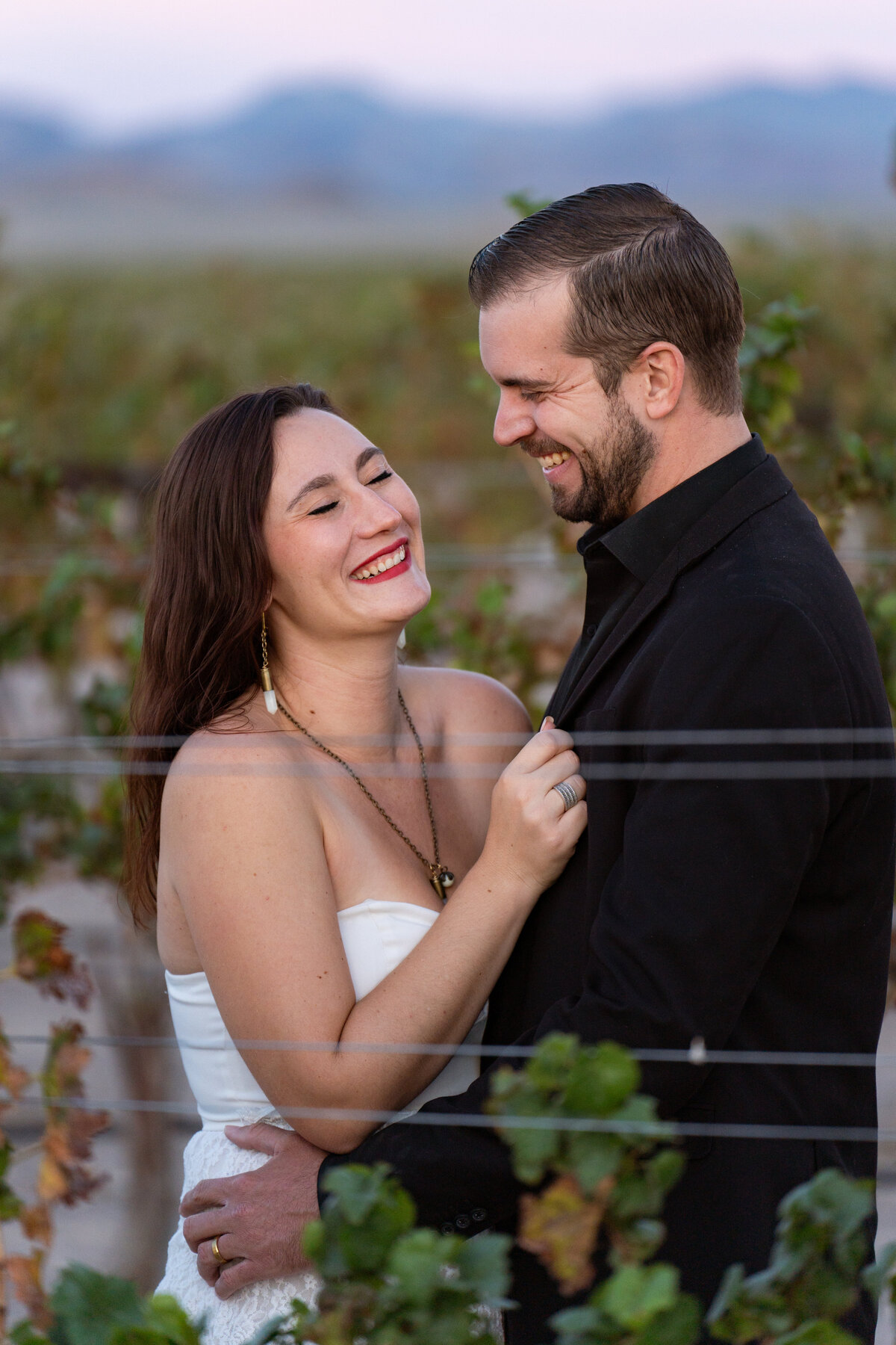 A couple laughing as they stand close together in a vineyard