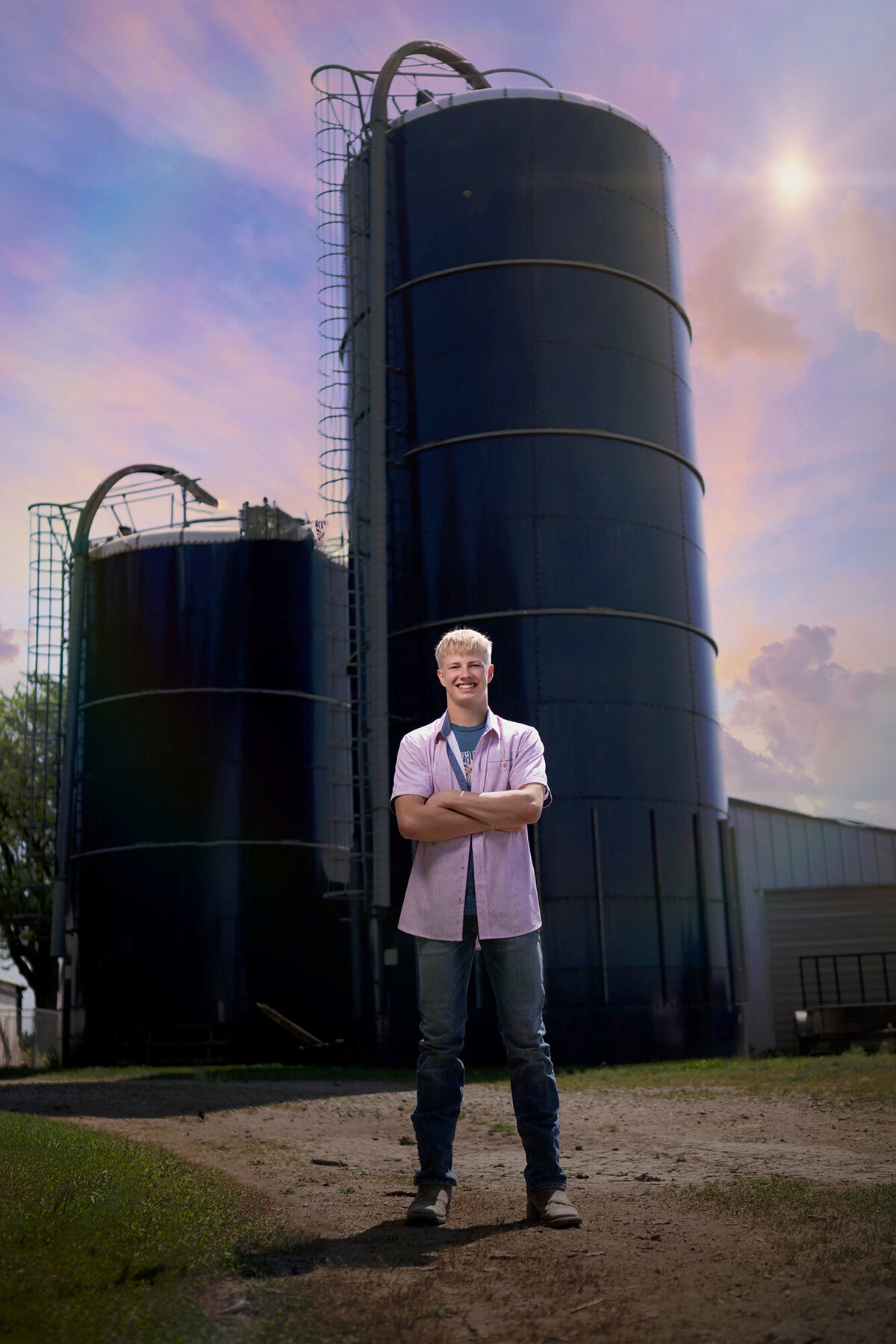 A Knoxville high school Iowa senior stands in a pink shirt in front of large farm silos