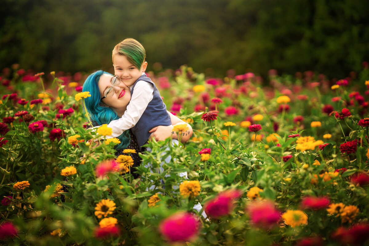 Mom and son are hugging in the zinnia fields.  Mom has blue hair and her son has green hair.  They are sitting in pink and yellow flowers  Both are smiling.