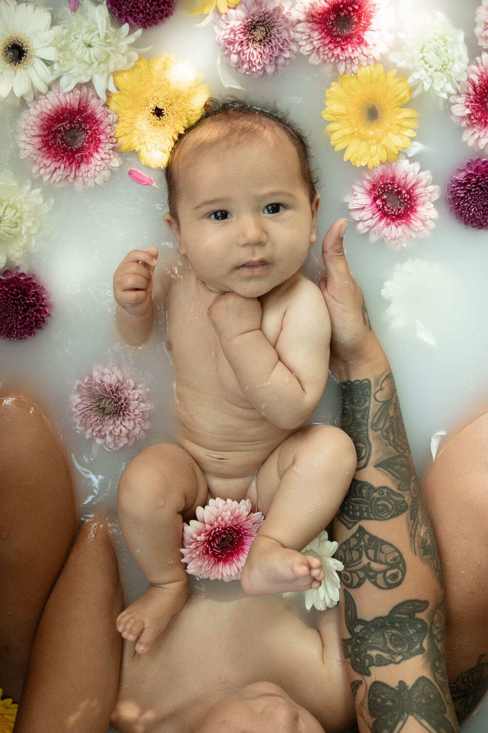 Close-up of a baby laying in a milk bath surrounded by flowers, looking at the camera