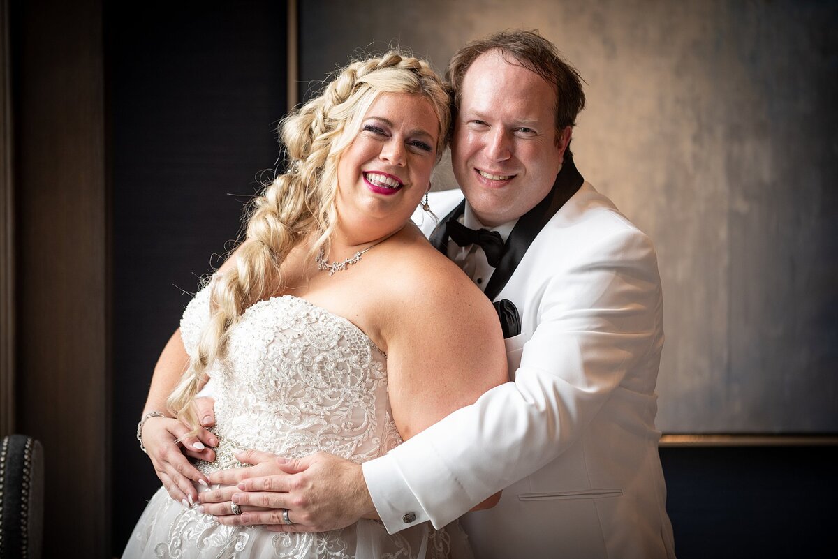 Plus sized bride in beaded strapless wedding gown with long blonde braid leans back and smiles at the camera as the groom, wearing a white tuxedo with a black shawl collar embraces her smiling at their Jewish wedding in Nashville