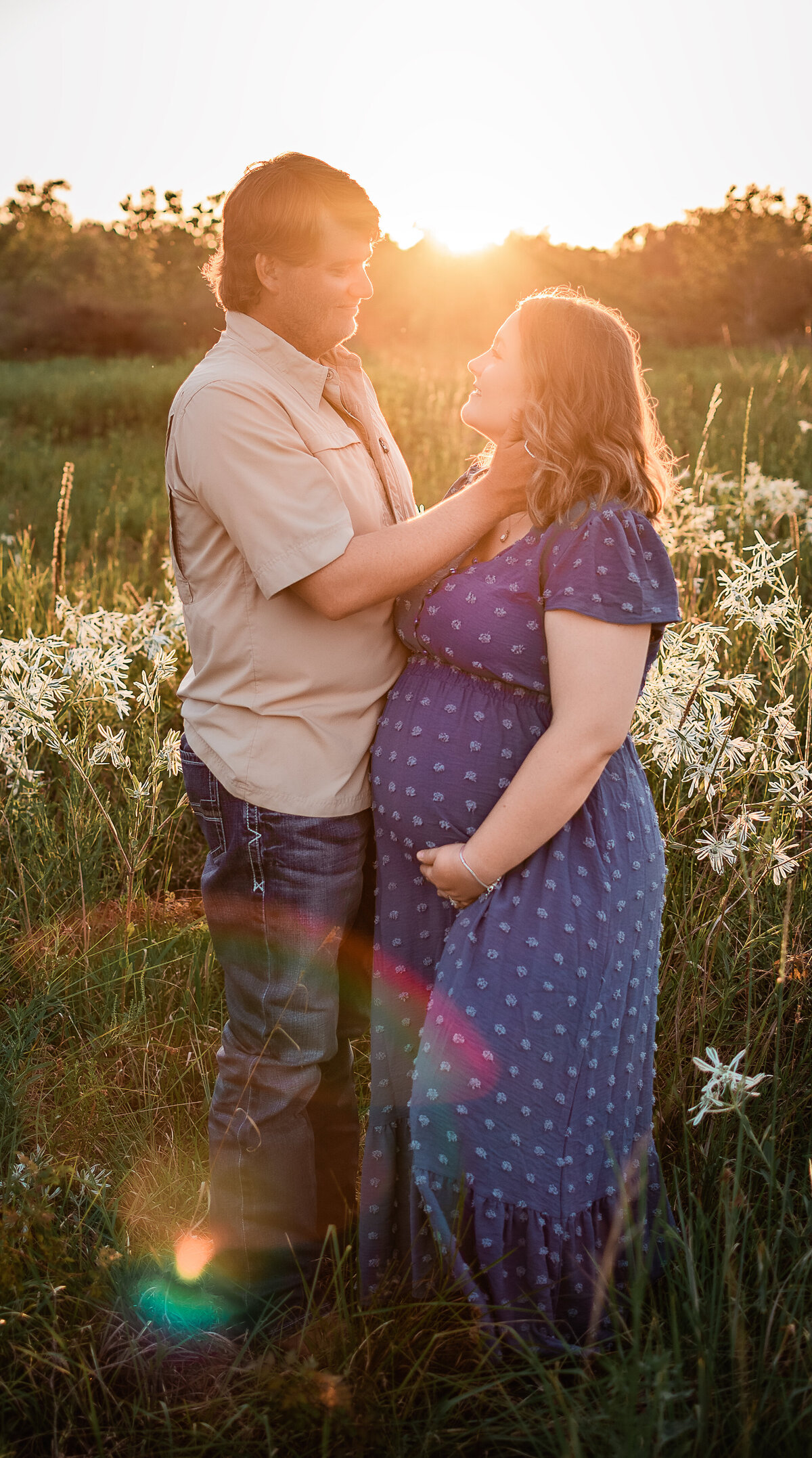 A mom and dad to be look at each other and smile in a field at sunset.