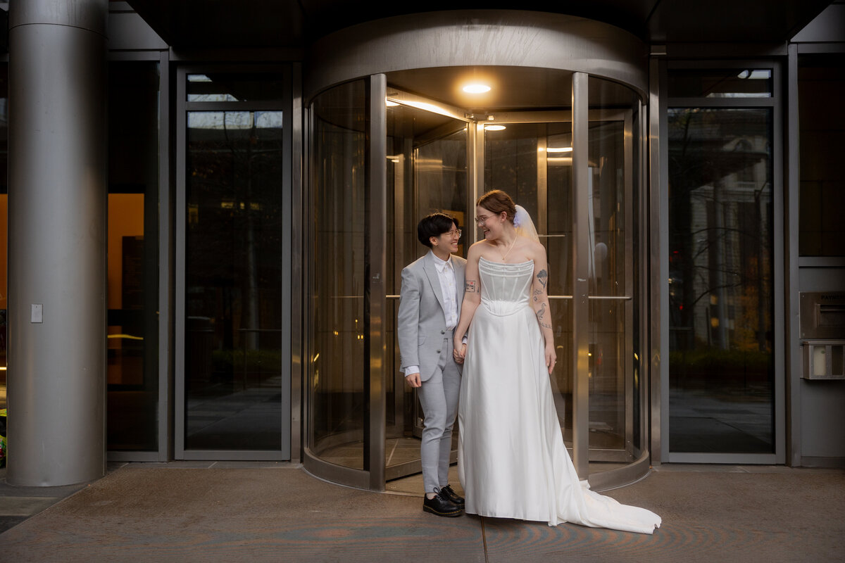 A stylish and memorable moment as the newlywed couple stands in front of the rotating door at the Seattle Courthouse, capturing the elegance and excitement of their wedding day.