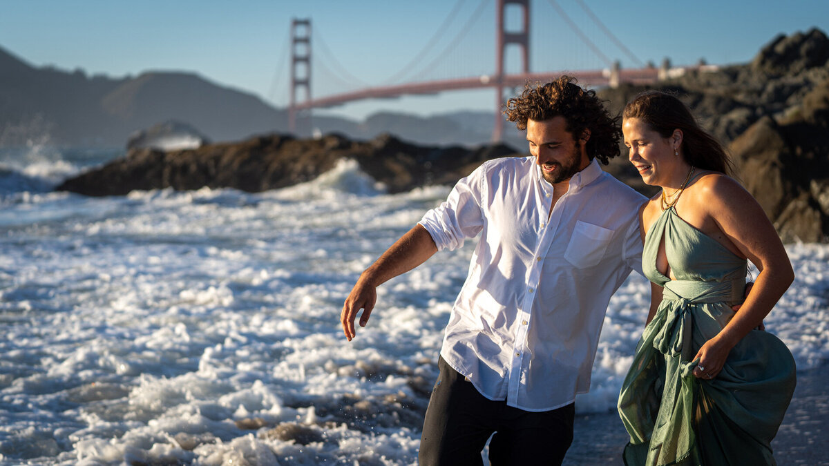 Couple holding hands while walking through the surf at the beach with the golden gate bridge in the background.