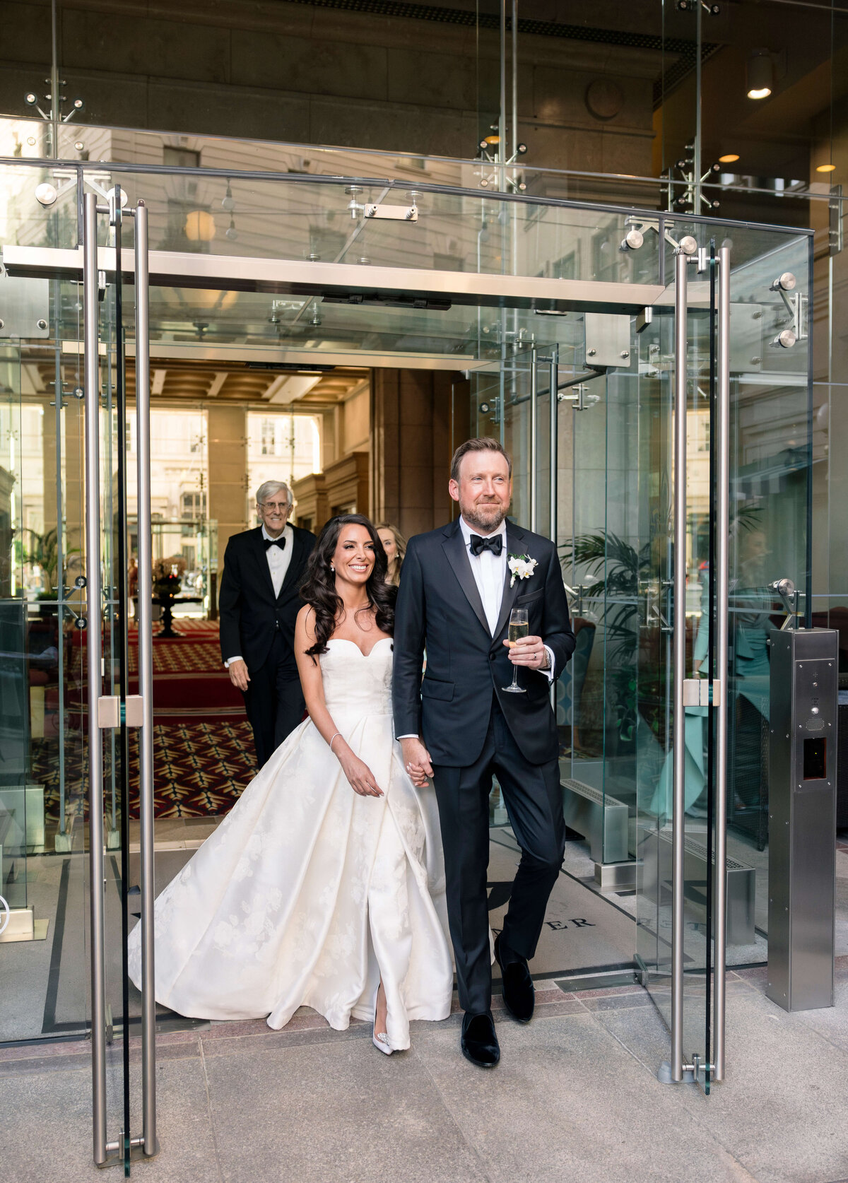 A bride and groom, dressed in formal wedding attire, walk hand in hand through glass doors. The bride wears a white gown, and the groom is in a tuxedo holding a drink. Two other people follow behind them, smiling.