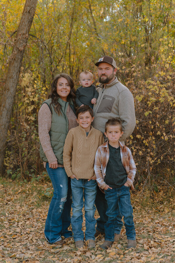 A family consisting of a mom, dad, and their 3 young sons pose standing together for a family photo. It's fall and the trees behind them are full of yellow leaves.