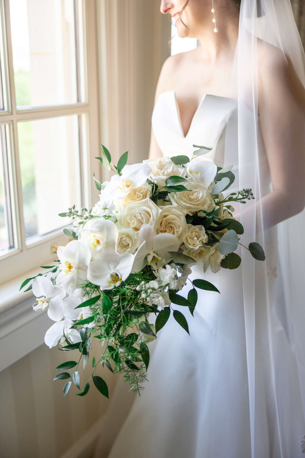 A bride in a white gown holds a cascading bouquet of white roses, orchids, and greenery. She stands near a window, with soft light highlighting her veil and the floral arrangement.
