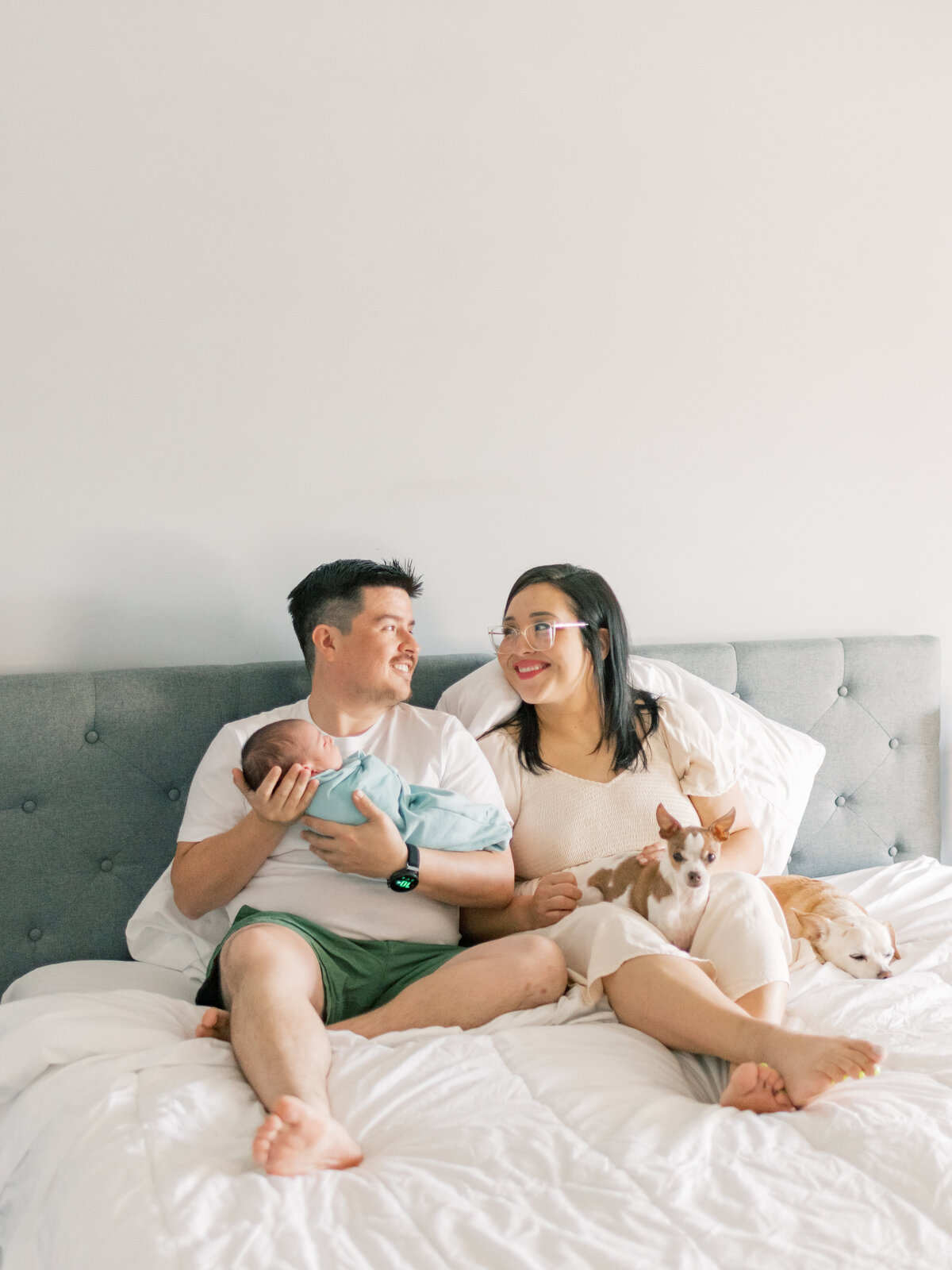 New parents sitting on their bed while holding their newborn baby with their two dogs.
