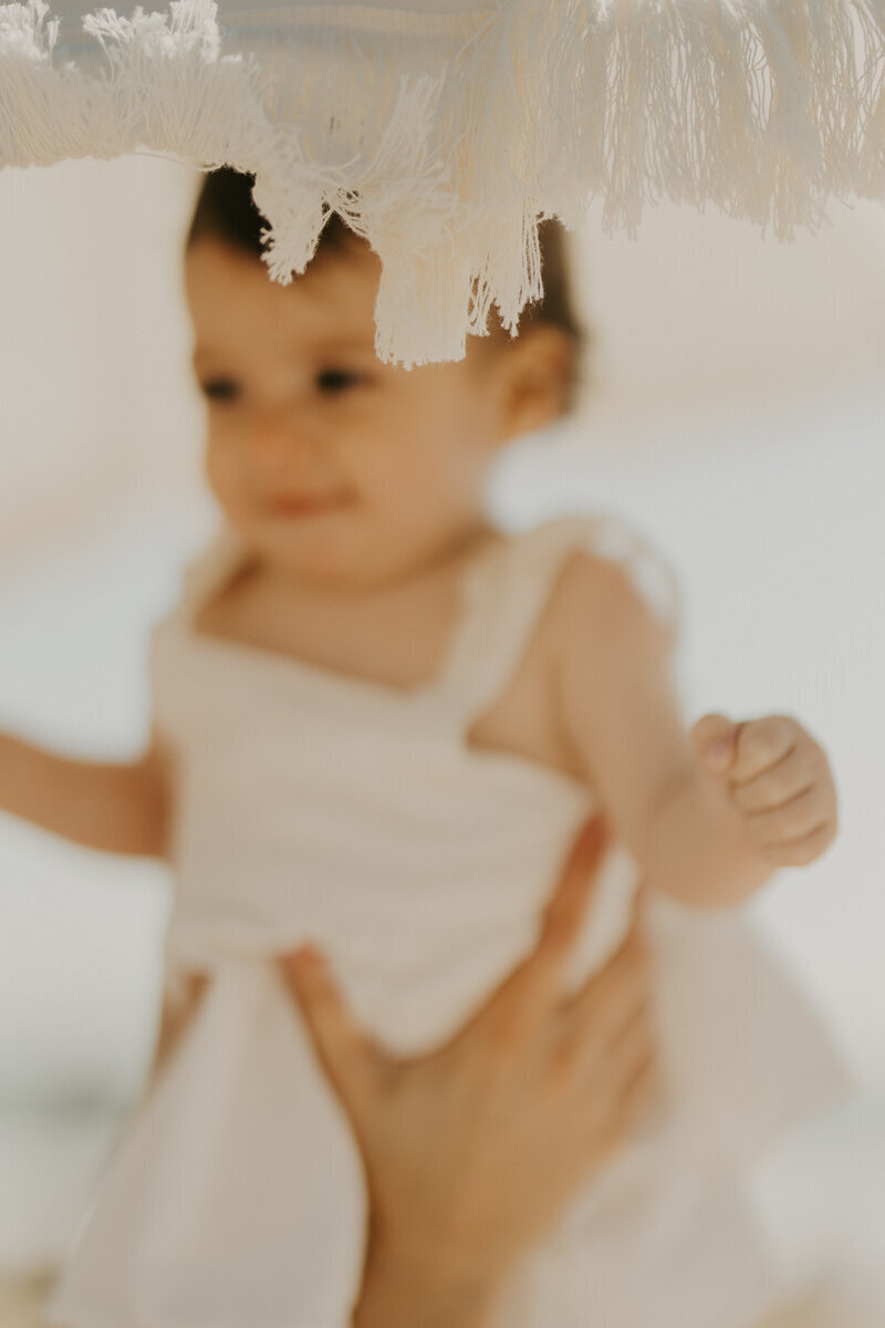 Petite fille en robe blanche floue portée à bout de bras sous une ombrelle capturée par Laura, photographe famille en Vendée.