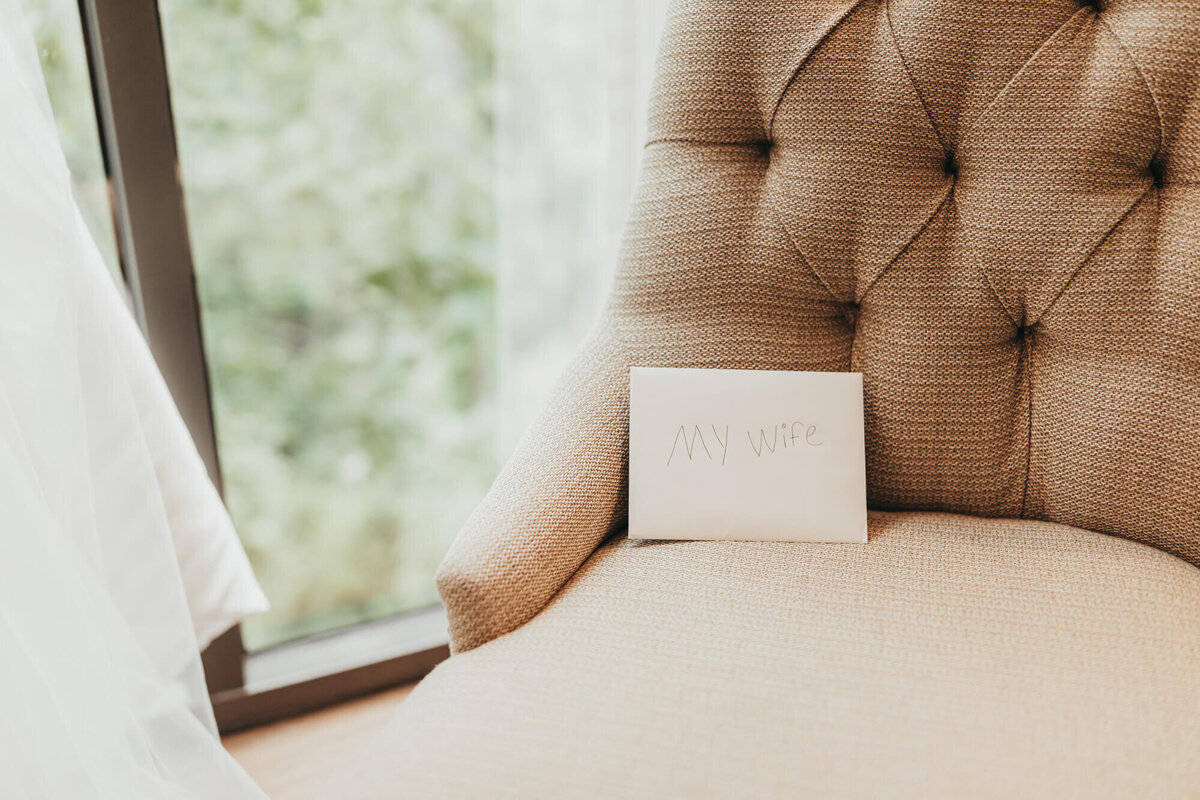 Card labeled "Wife" sits on chair against wall of windows.