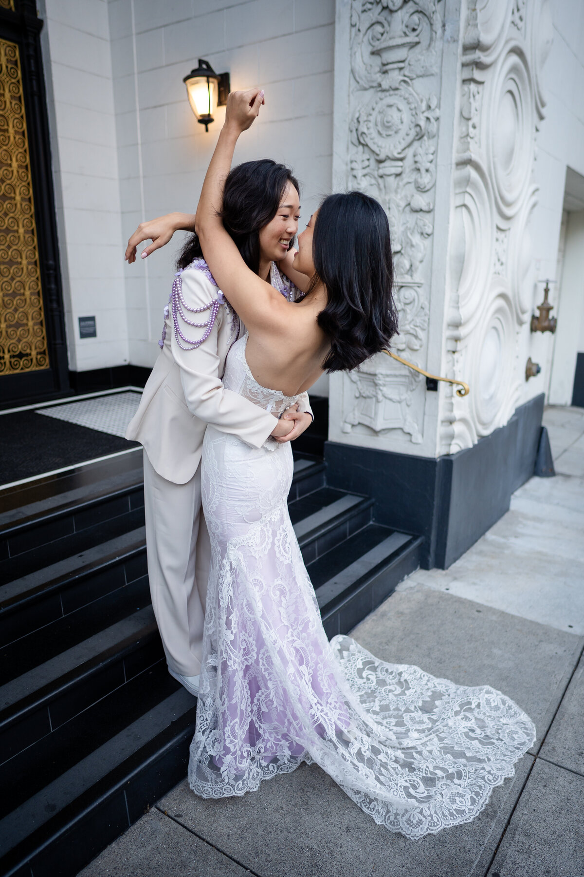 Queer lesbian couple in San Francisco during dawn embracing each other on a art deco inspired staircase with one wearing colored Quill Claire Pettibone gown