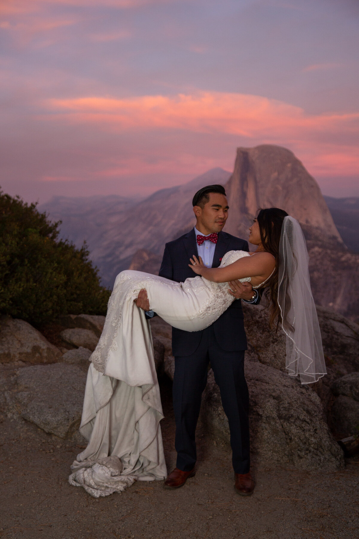 A groom stands holding his bride in his arms as her veil hangs behind her and the sunsets making the sky a beautiful pink color.