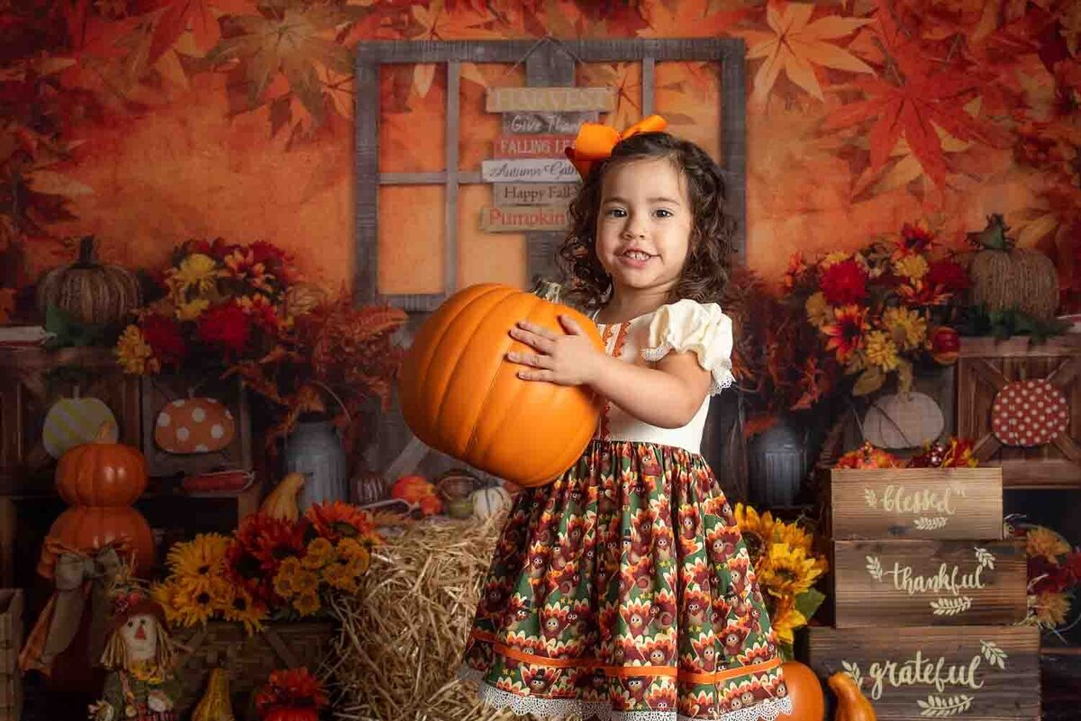 Toddler girl holding pumpkin in festive fall studio milestone set with autumn leaves and seasonal décor in Birmingham photography studio.