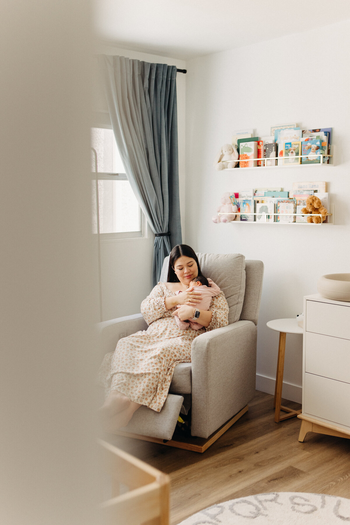 Mom sits in rocking chair in the baby nursery holding sleeping newborn.