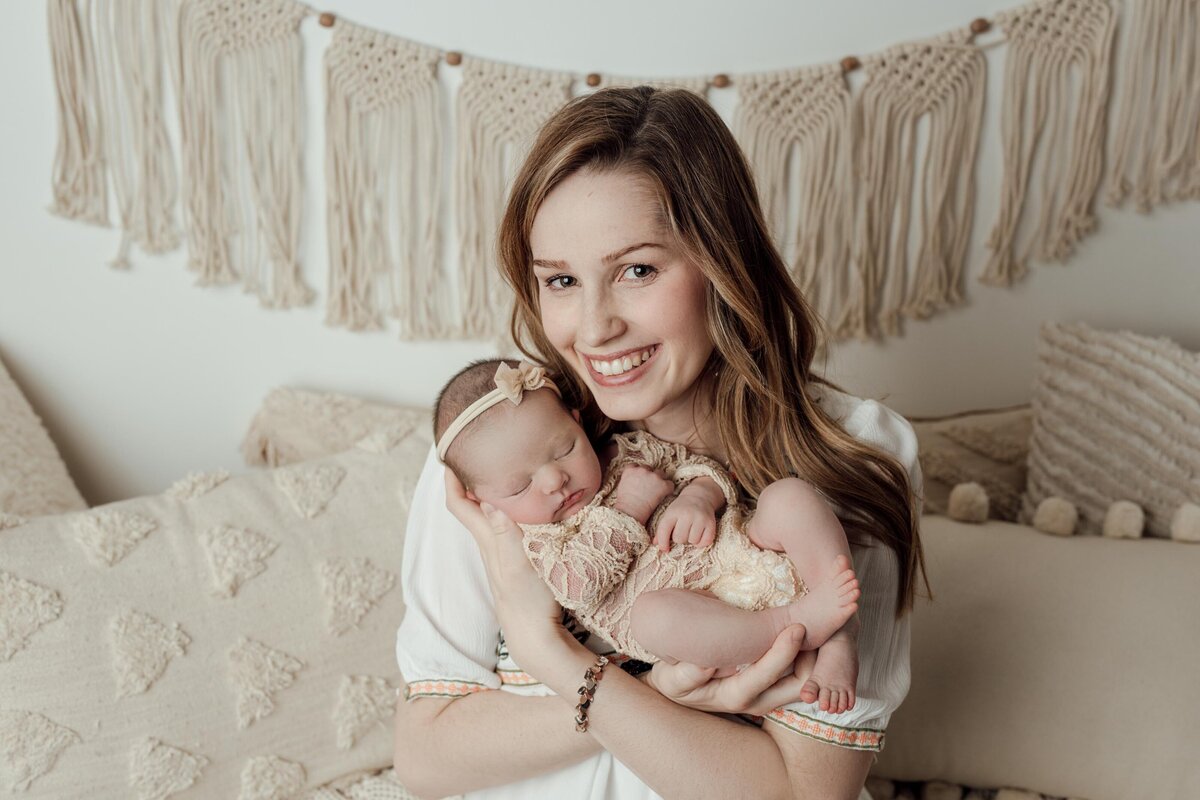 Studio newborn photography - Mom in studio lifestyle session holding baby under her chin. Baby is in a cream bodysuit with matching headband. Mom is smiling at the camera. Boho fringe in the background.